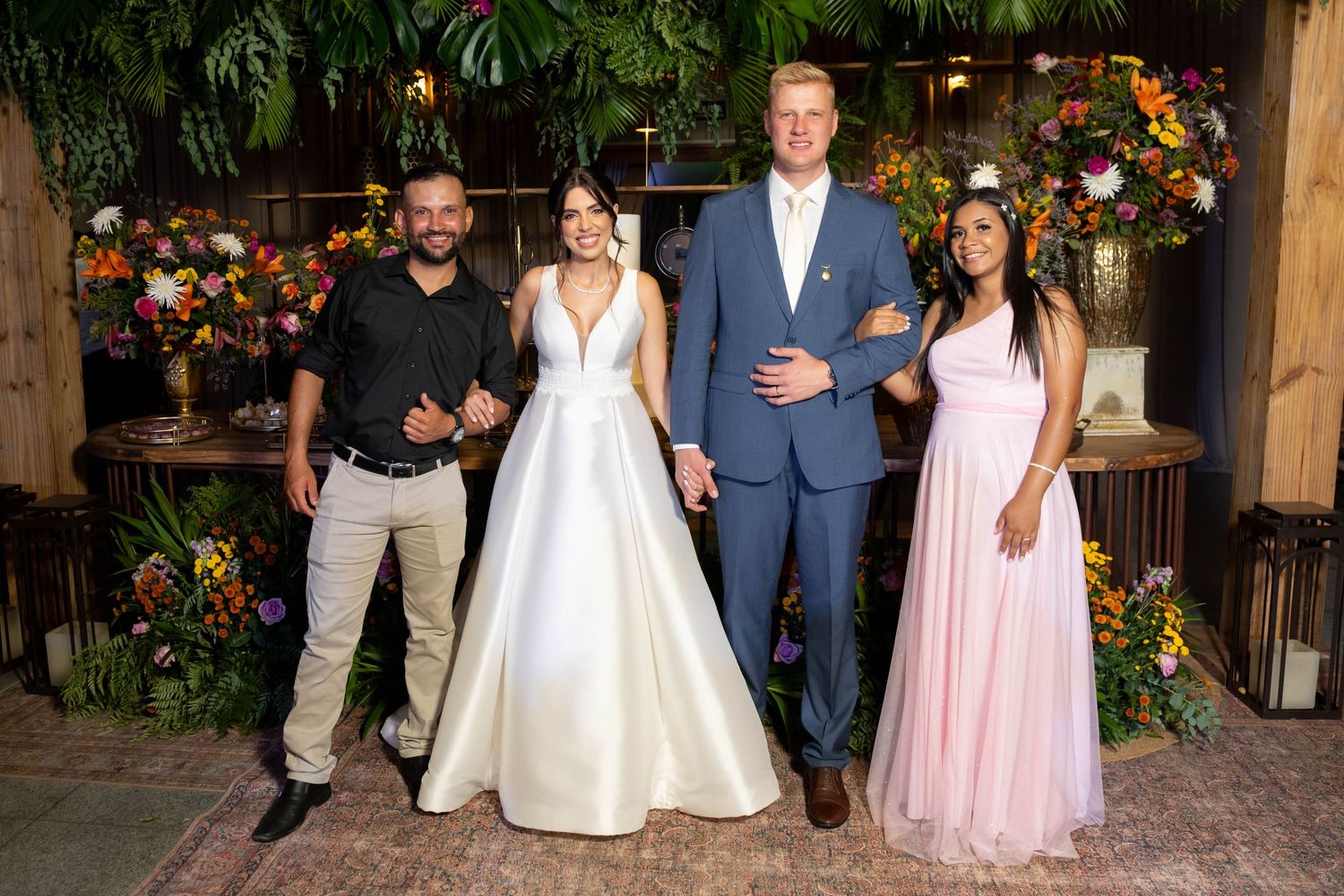 Two couples pose at a wedding, bride in white gown, groom in blue suit, guests in black and pink formal attire.