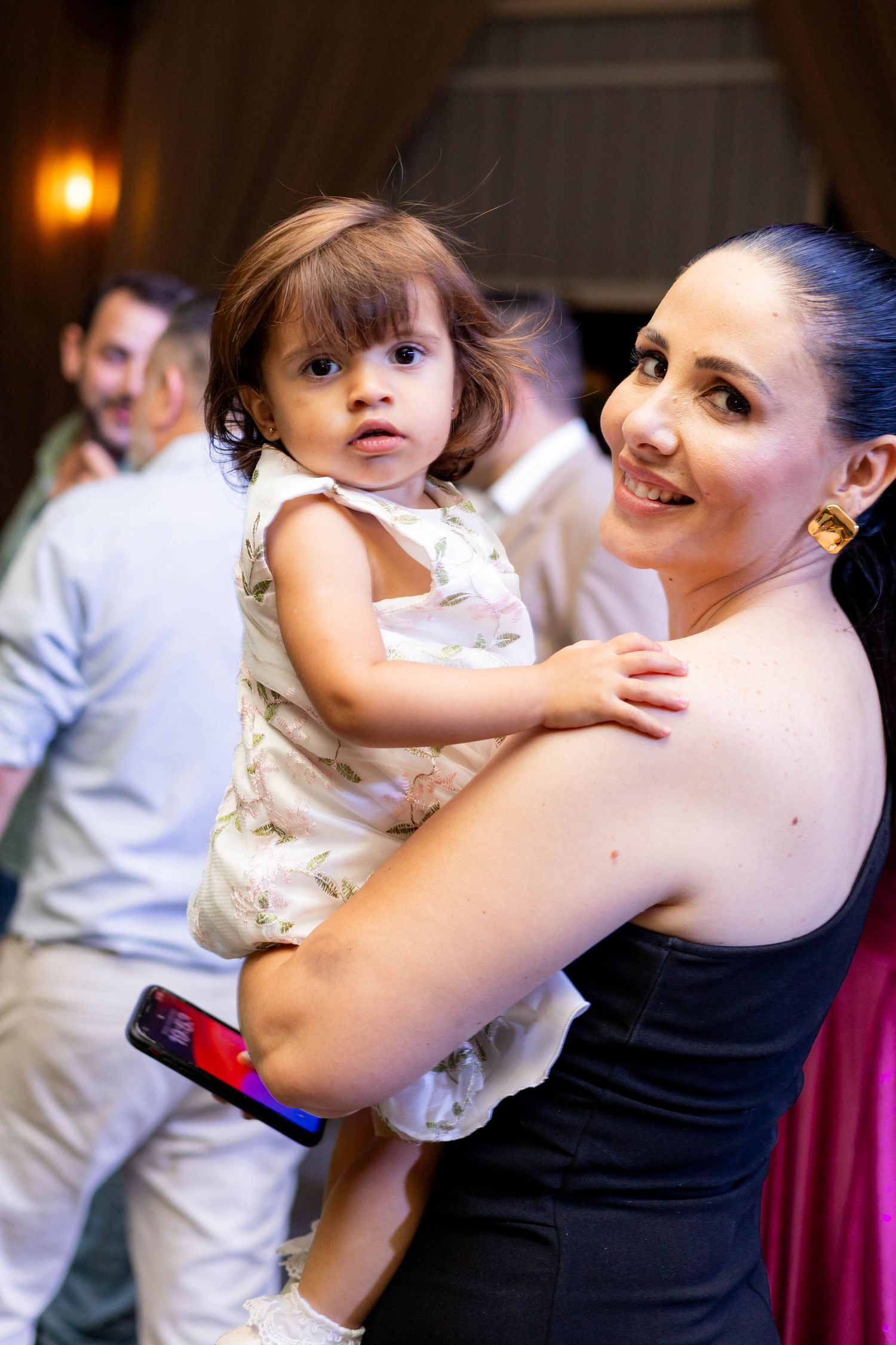 A smiling woman in a black dress holds a cute toddler girl in a white dress at a party event.