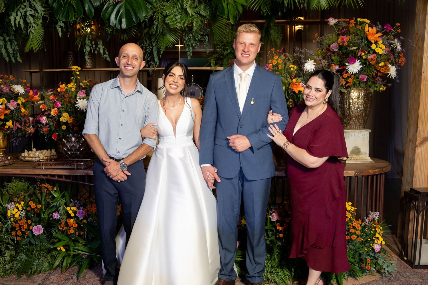 Wedding couple in white gown and blue suit pose with two guests in front of lush floral backdrop.