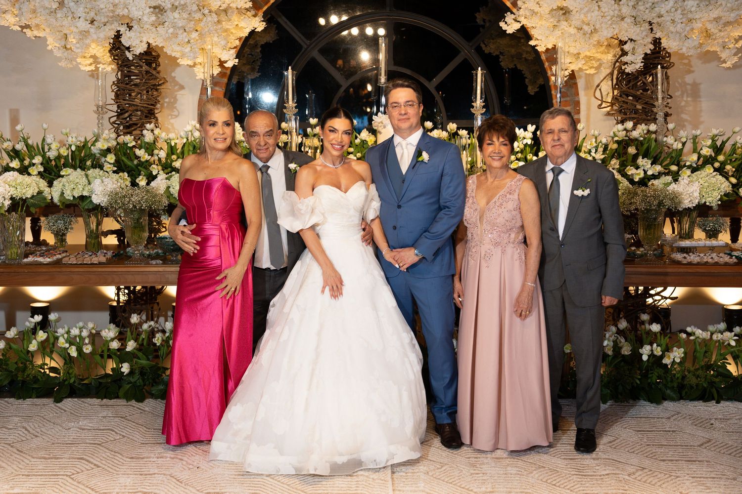 Wedding party of six posing elegantly in formal attire, bride in white gown, groom in blue suit, at floral reception.