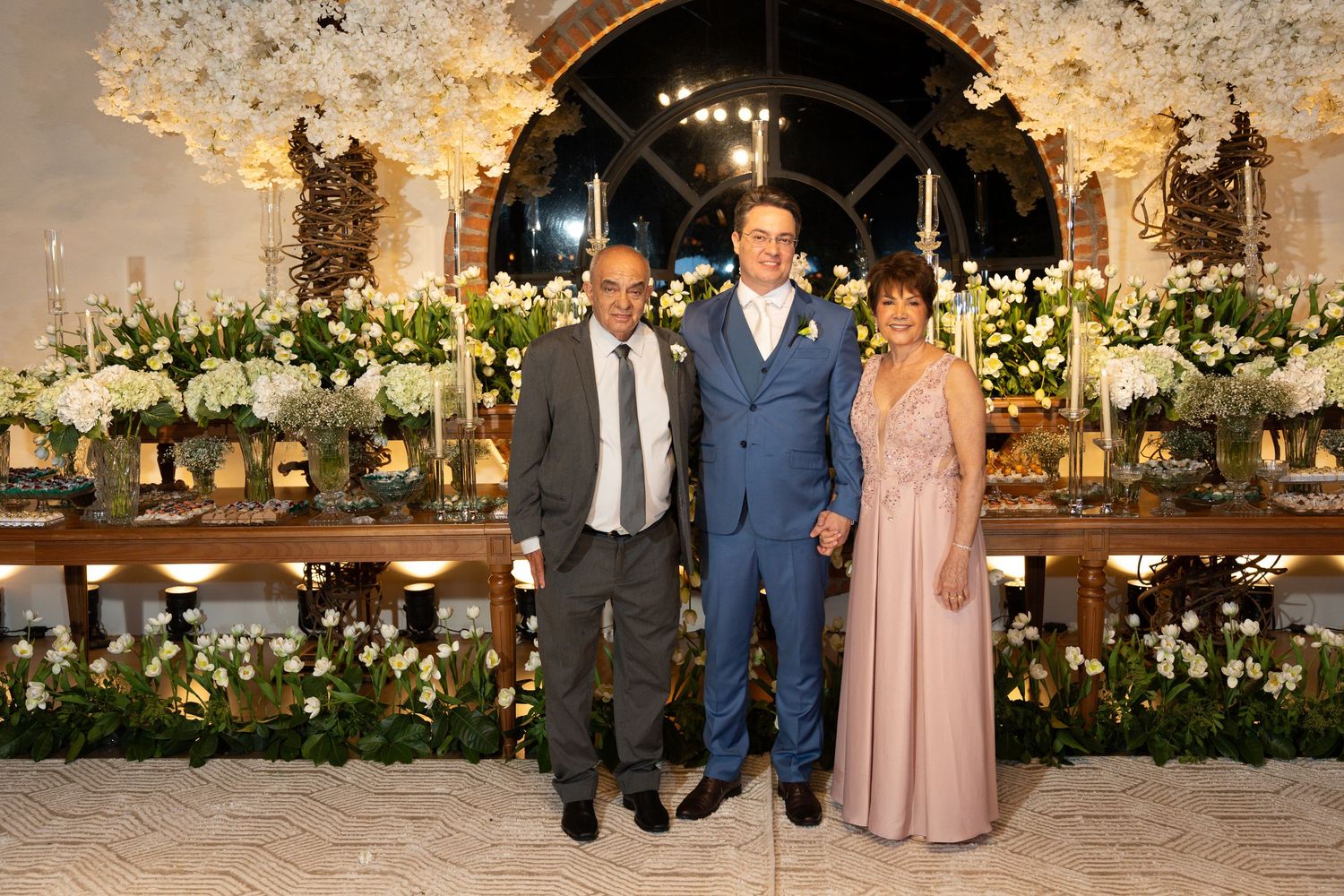 Three elegantly dressed people posing together at a floral wedding reception with white flower arrangements.