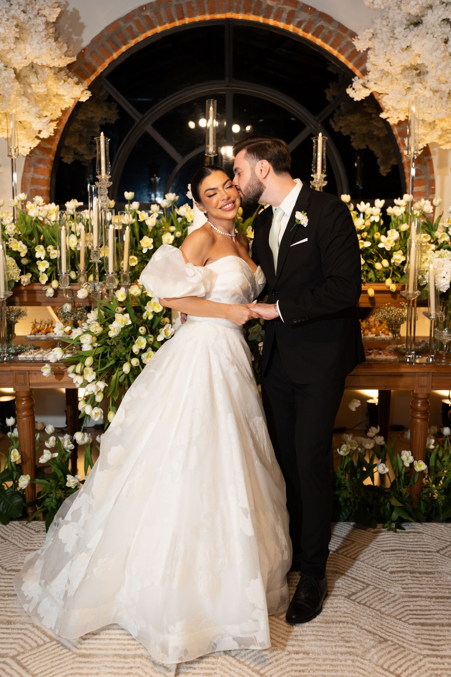 Bride in white ball gown and groom in black tuxedo embrace at elegant wedding reception with floral backdrop.