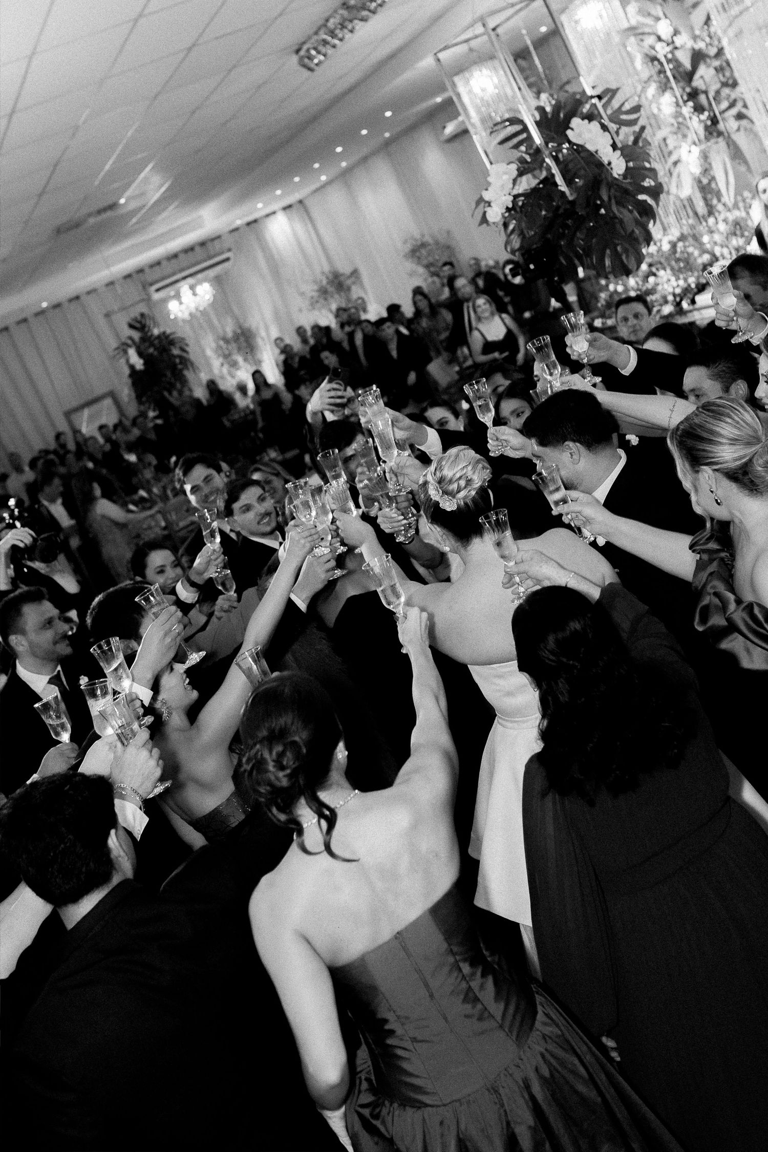 Black and white photo of wedding guests raising glasses in a champagne toast at a reception.