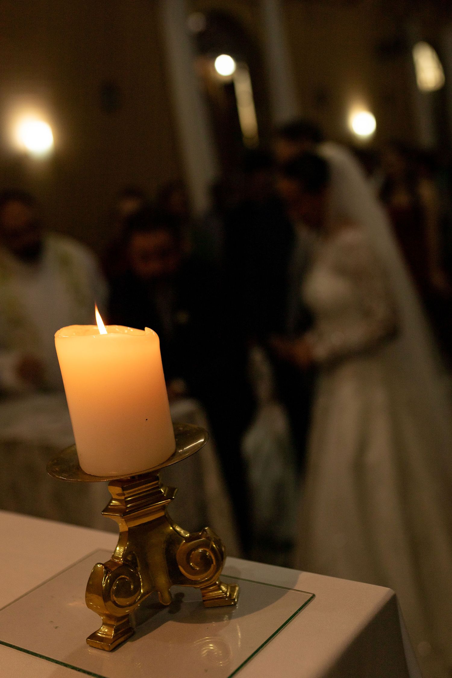 A lit pillar candle in a gold holder with blurred bride and groom exchanging vows in the background.