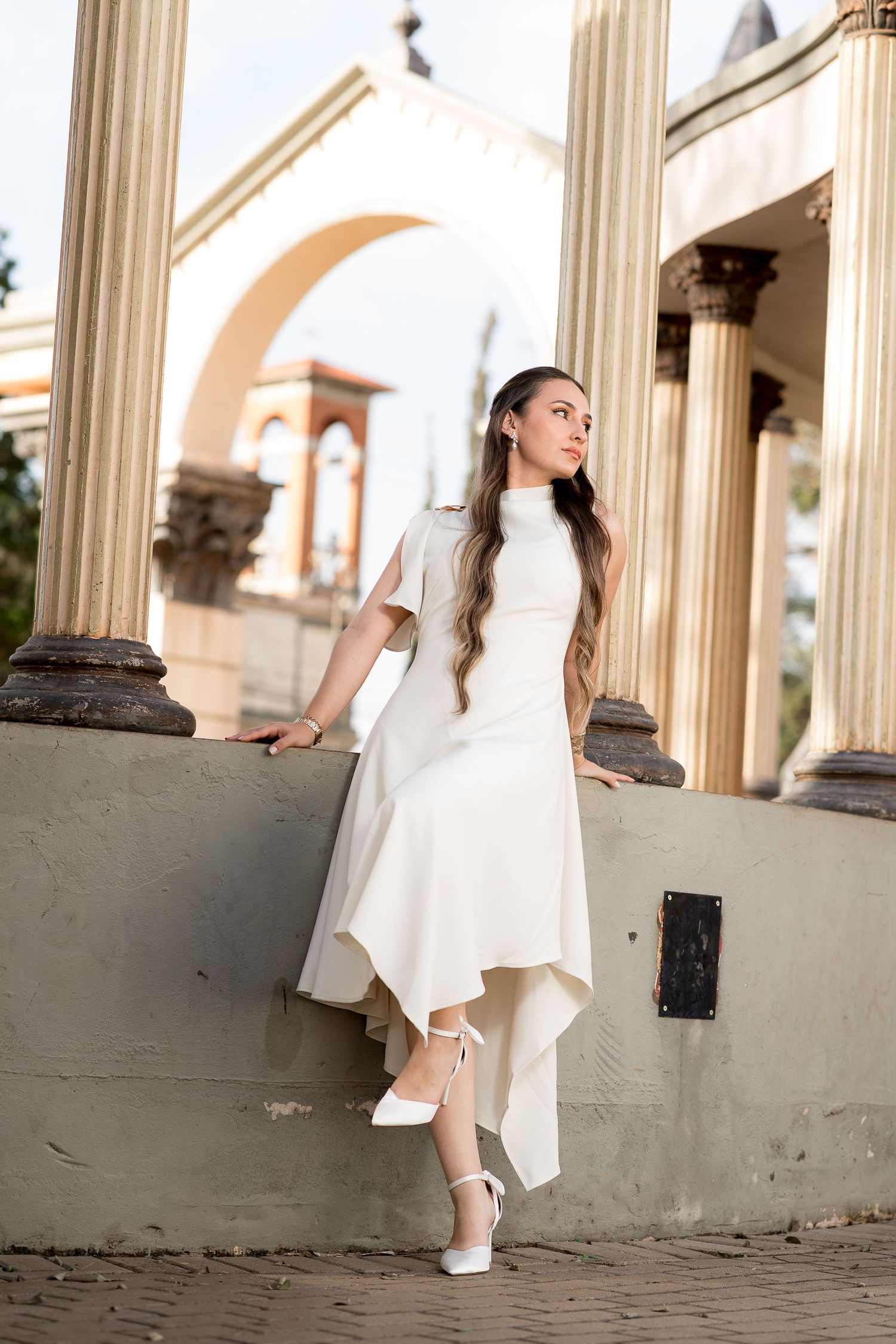Woman in elegant white dress and heels poses gracefully between classical columns at an ornate outdoor venue.