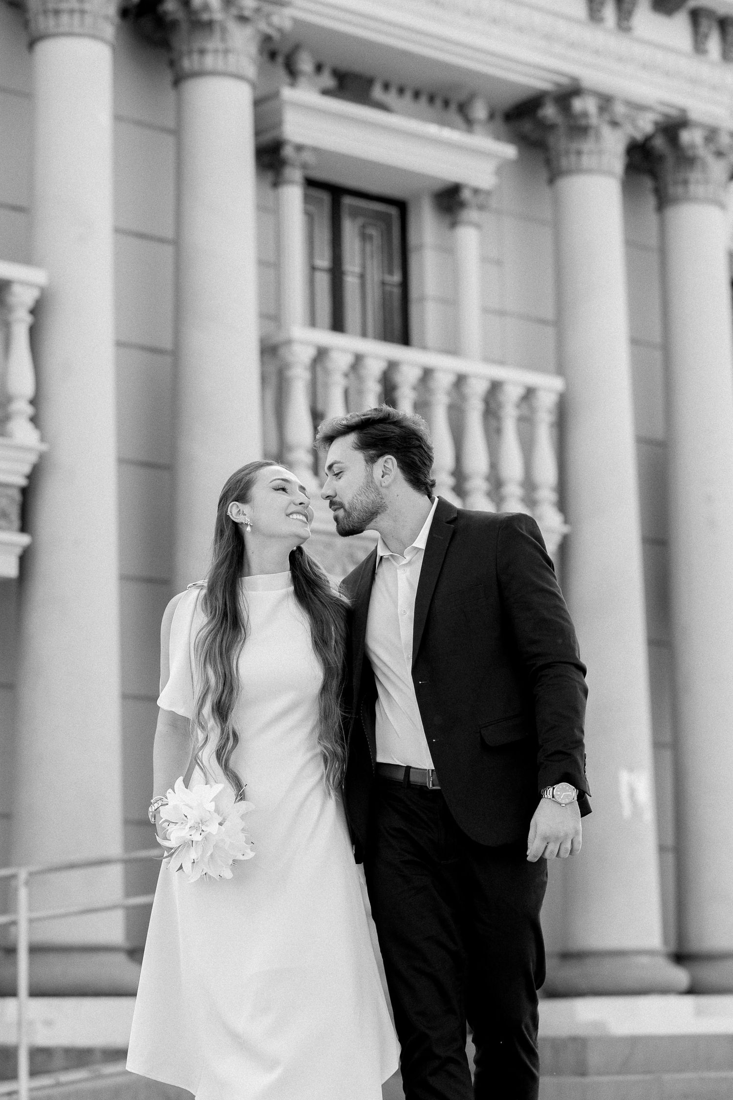 Black and white photo of a bride and groom sharing a tender moment on steps of a classical building with columns.