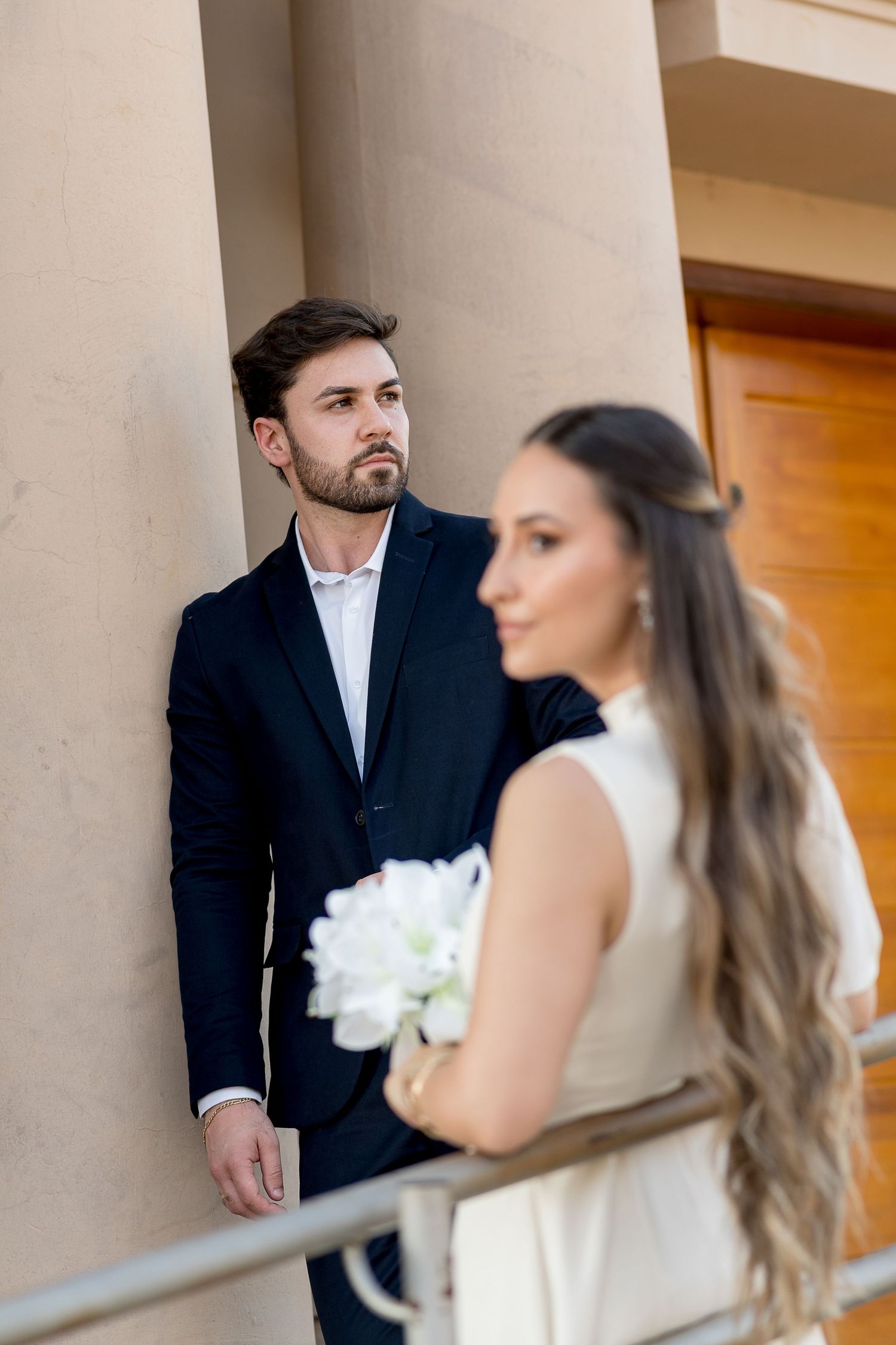 Groom in dark suit leans against column while bride in white dress holds bouquet in foreground.