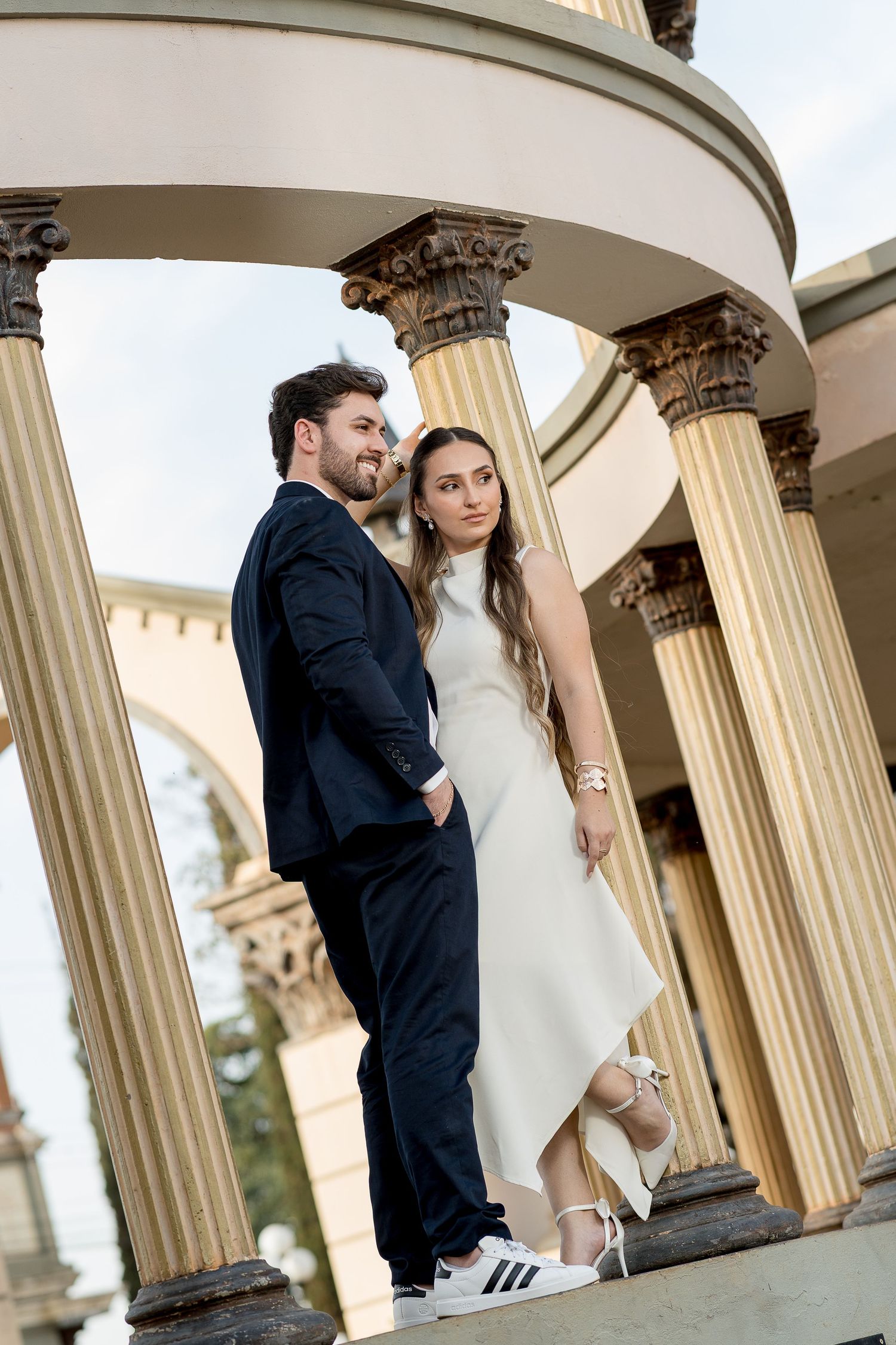 Elegant couple posing between classical columns, man in navy suit, woman in white midi dress with Adidas sneakers.