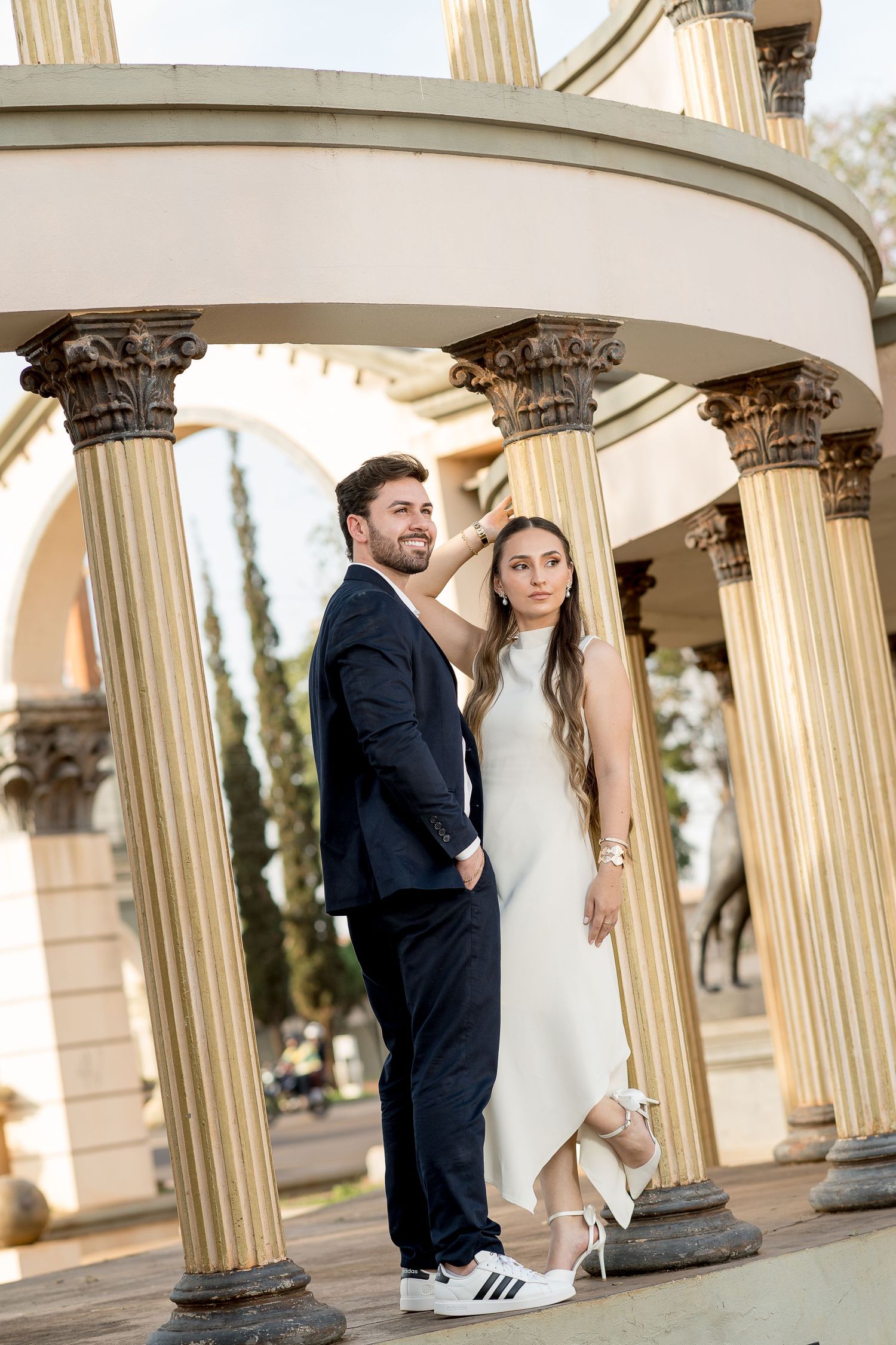 Couple posing elegantly among classical columns, woman in white dress with sneakers, man in dark suit, outdoor setting.