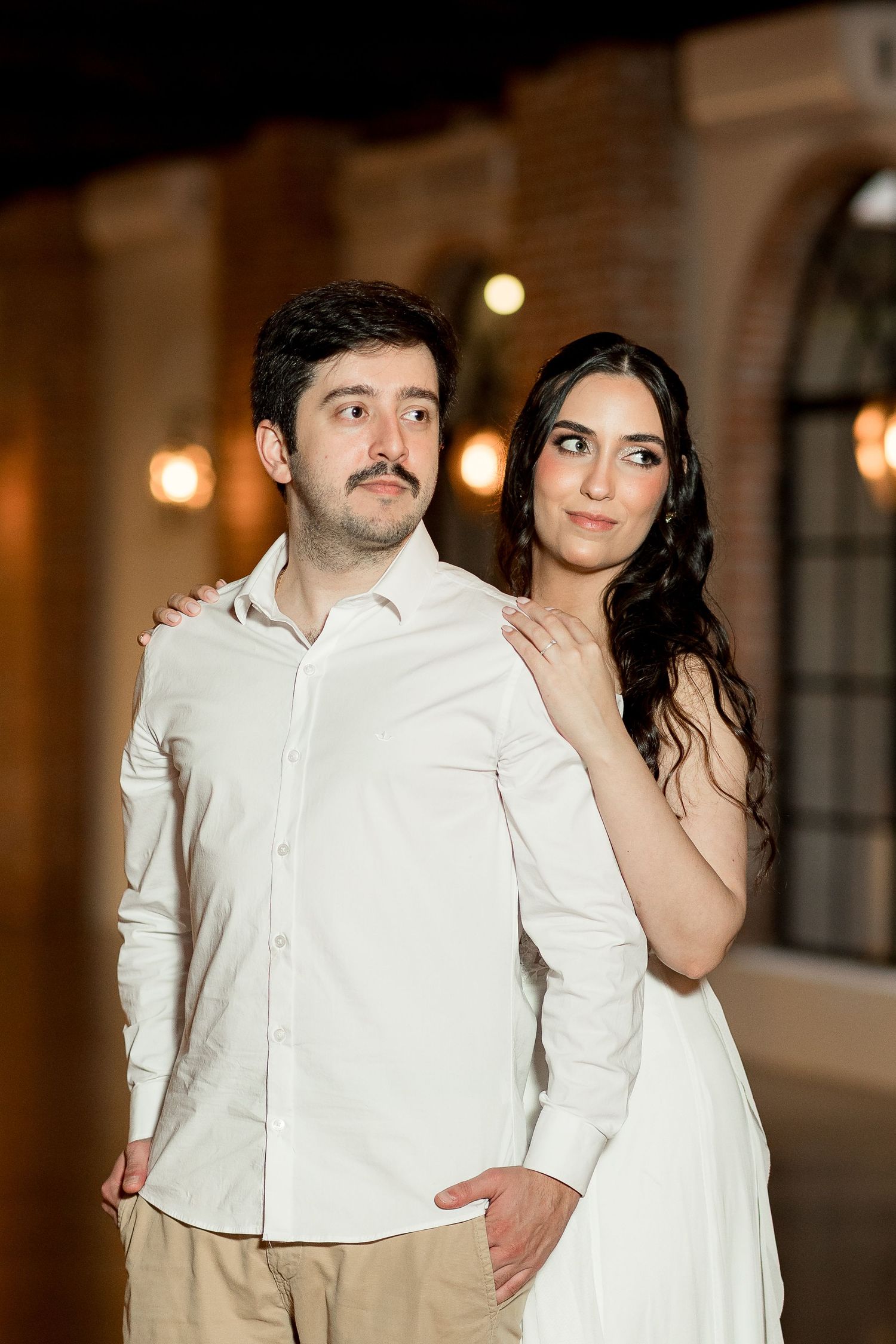 Couple in white outfits posing at night, woman resting hand on mans shoulder near arched windows with warm lights.