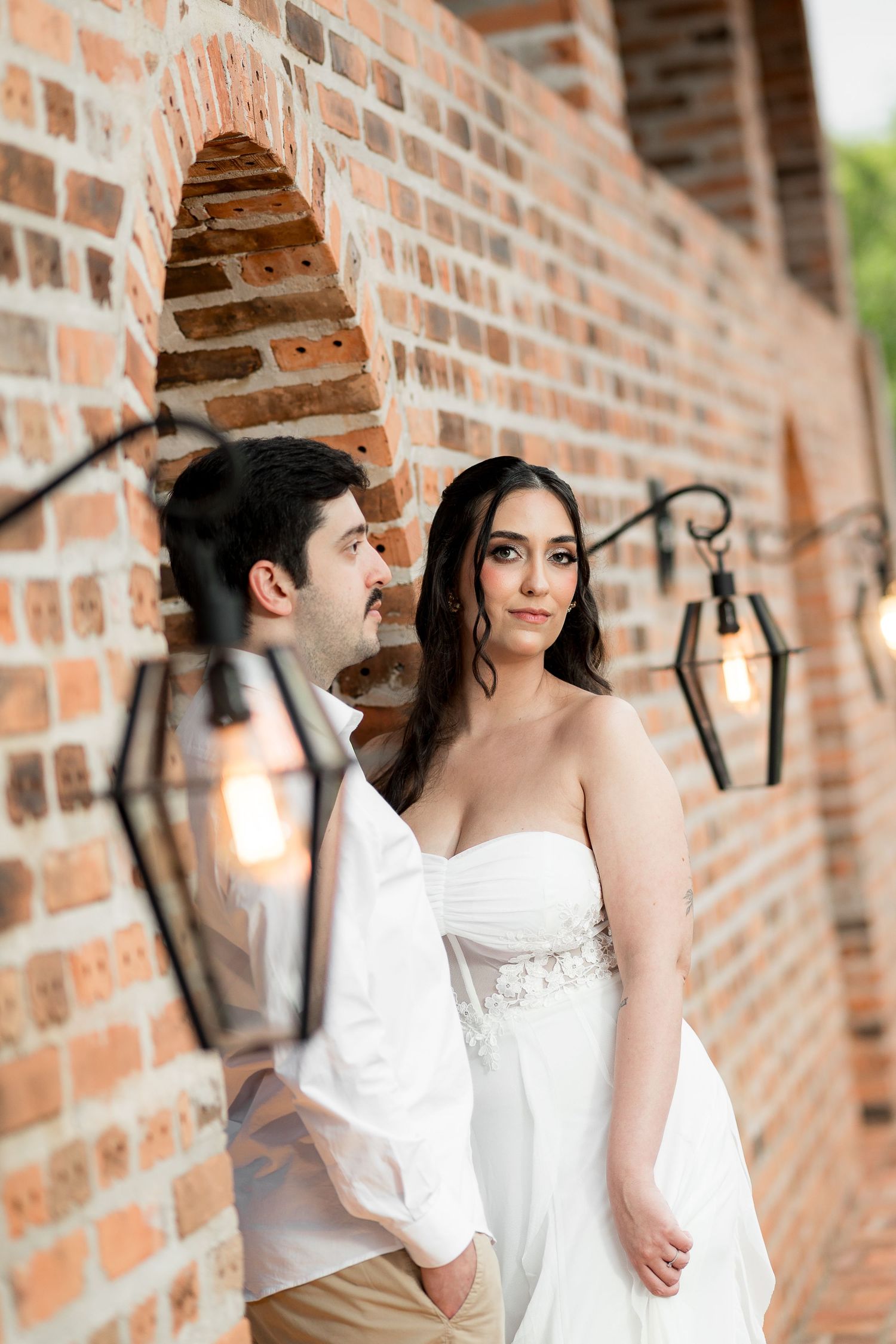A bride in a white strapless gown and groom in white shirt pose by a rustic brick wall with vintage lanterns.