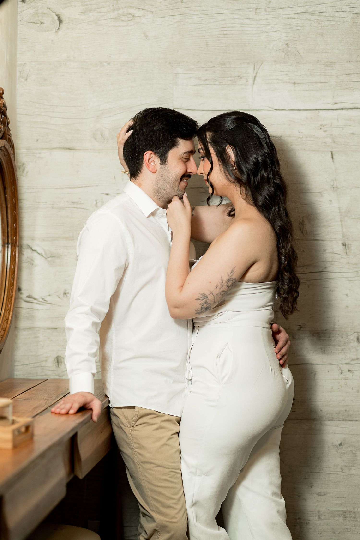 A romantic couple in white wedding attire sharing an intimate moment, leaning close together against a light wood wall.