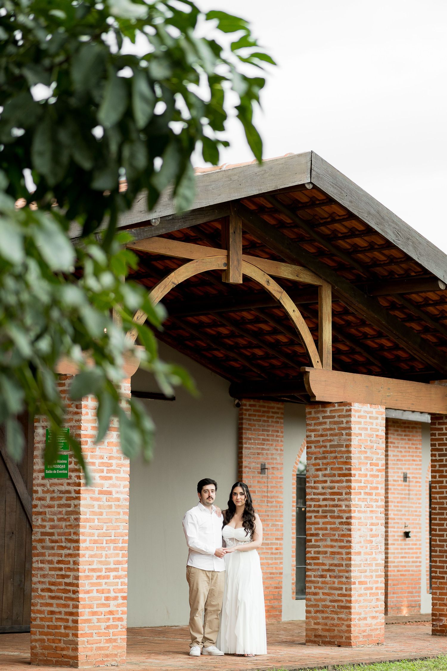 Couple in white outfits standing under a wooden pergola with brick columns surrounded by lush greenery.