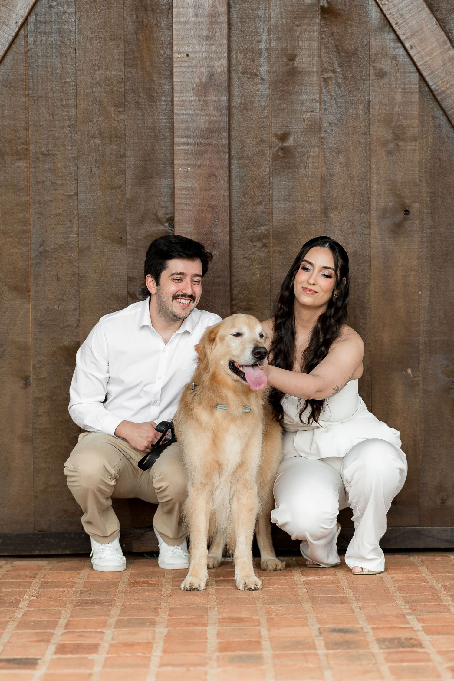 Smiling couple in white outfits crouching beside a golden retriever dog in front of a rustic wooden barn door.
