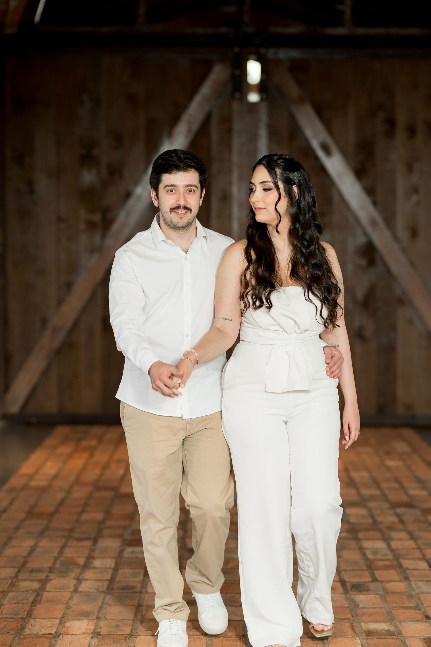 Couple in white outfits holding hands in a rustic barn with wooden walls and brick flooring.
