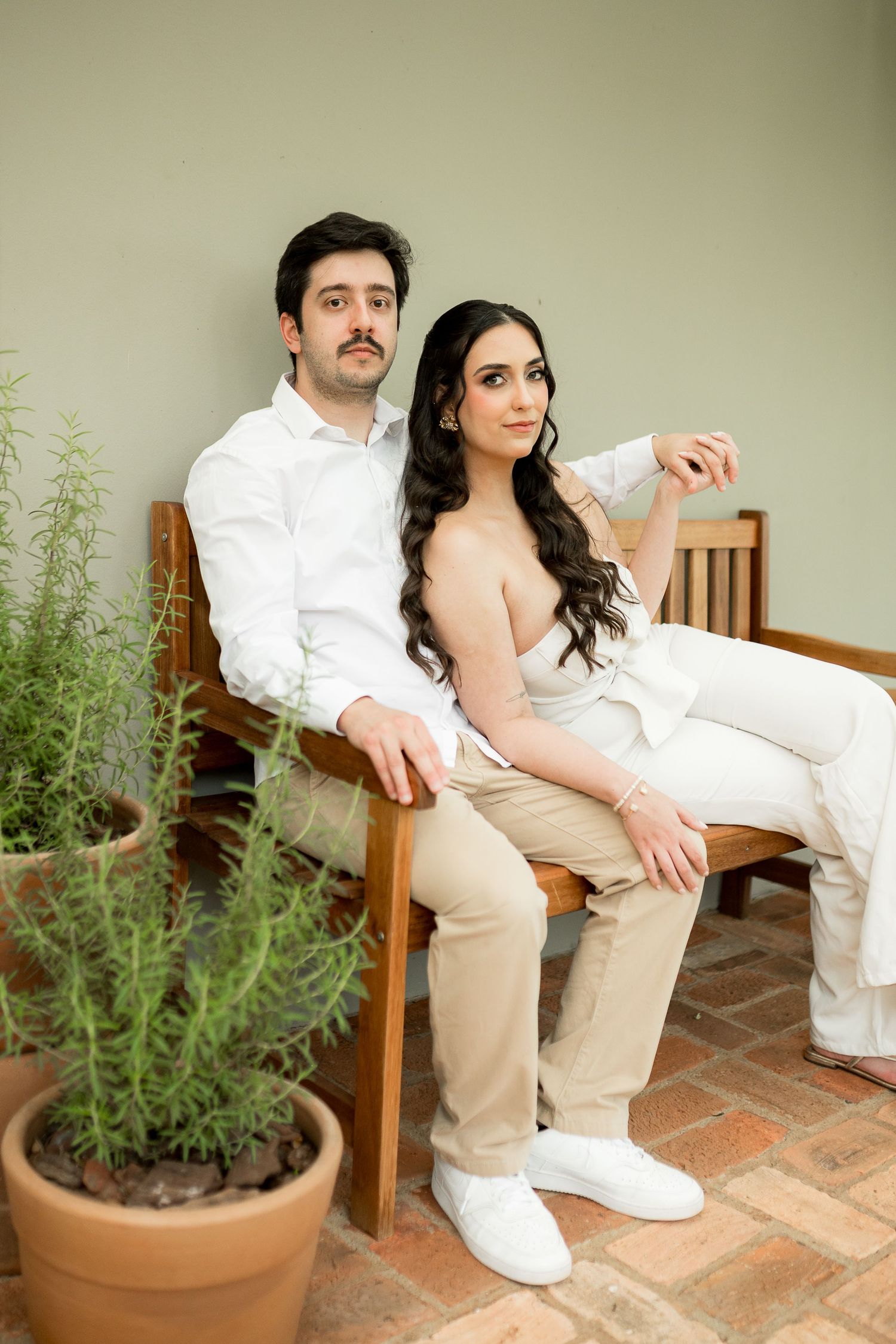 Couple in matching beige and white outfits sitting on a wooden bench near potted plants against a sage green wall.