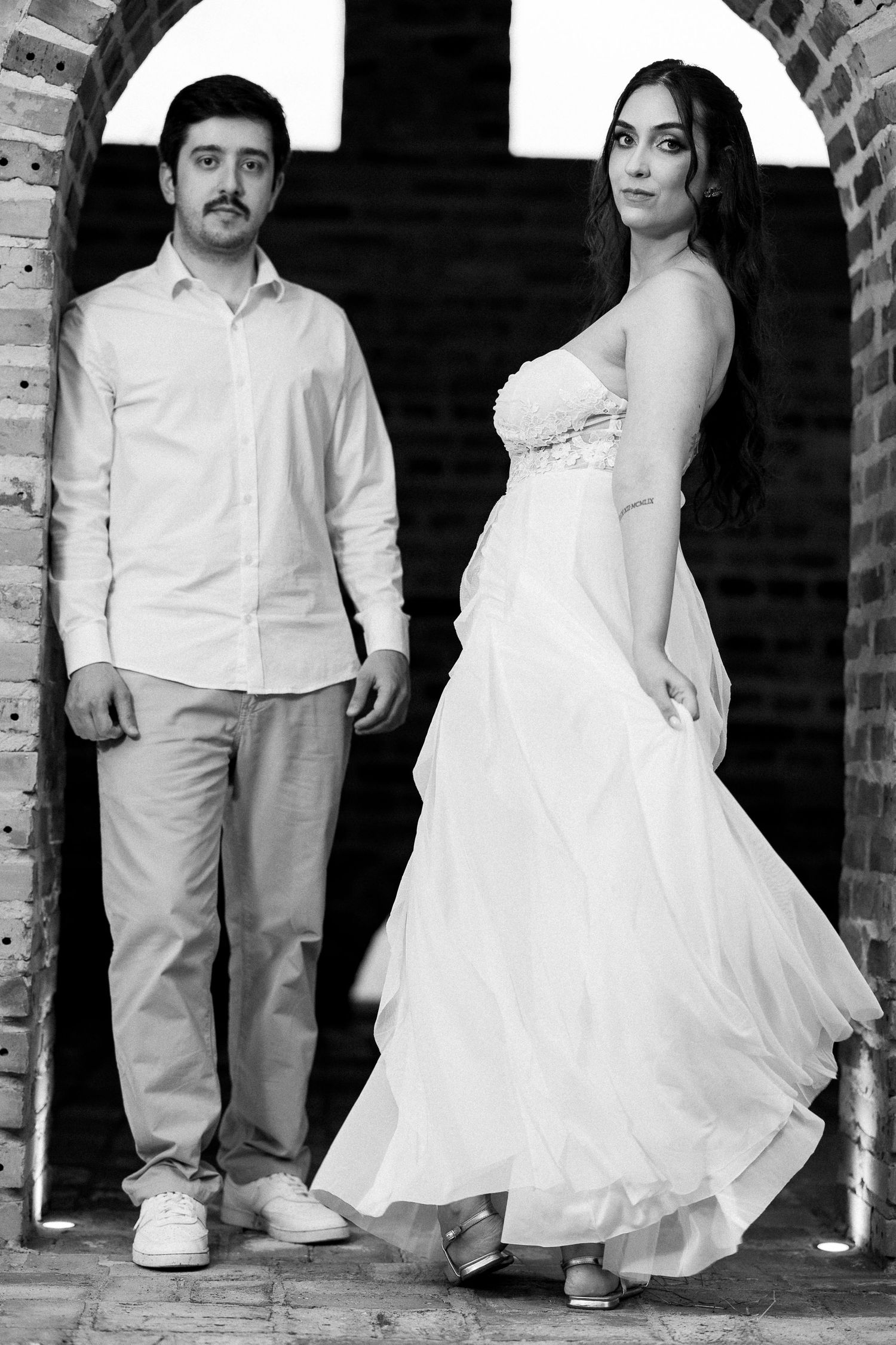 Black and white photo of bride and groom posing in brick archway, bride in elegant white dress.