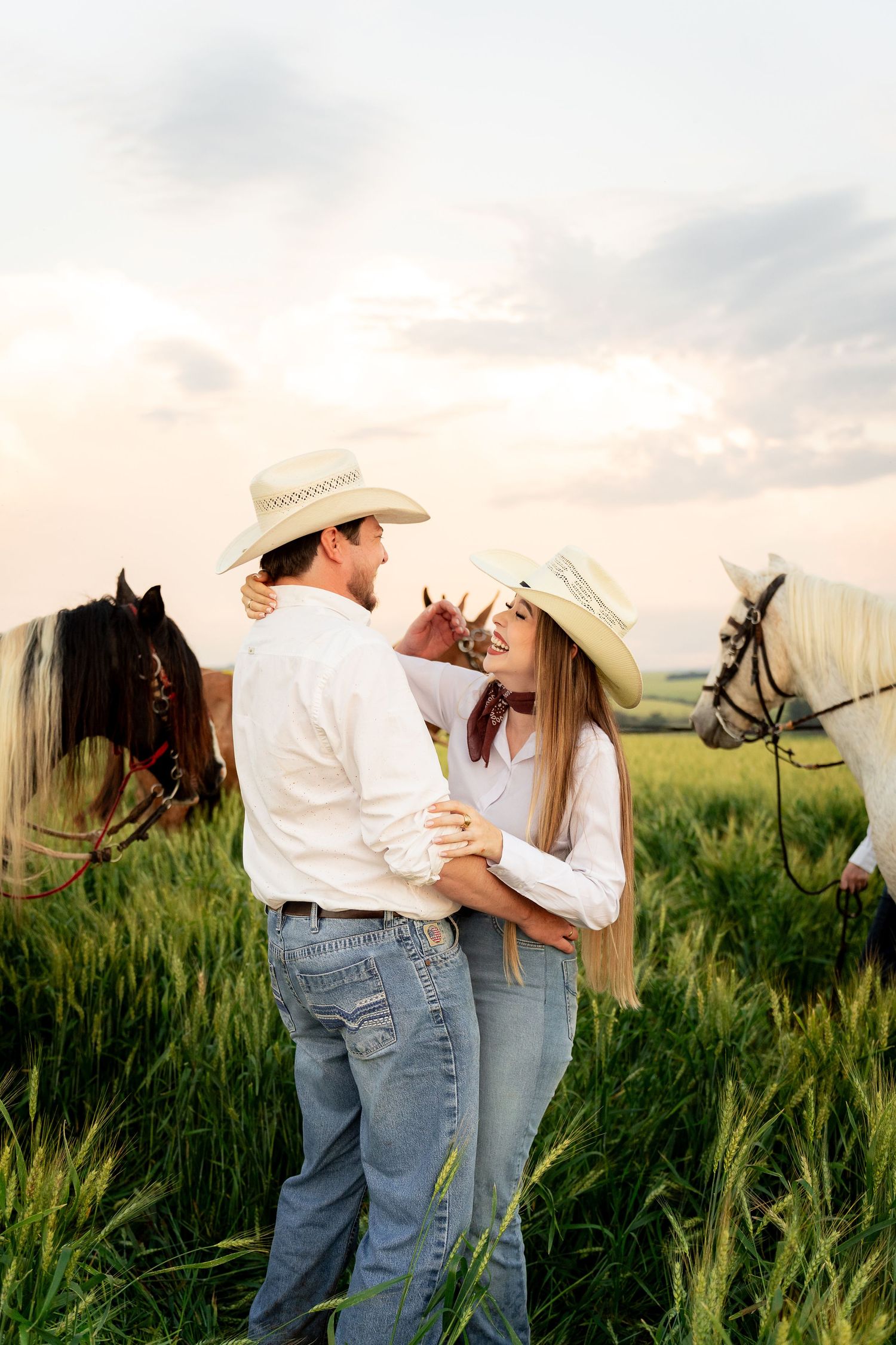 Couple in cowboy hats embracing in a green field with brown and white horses in the background at sunset.
