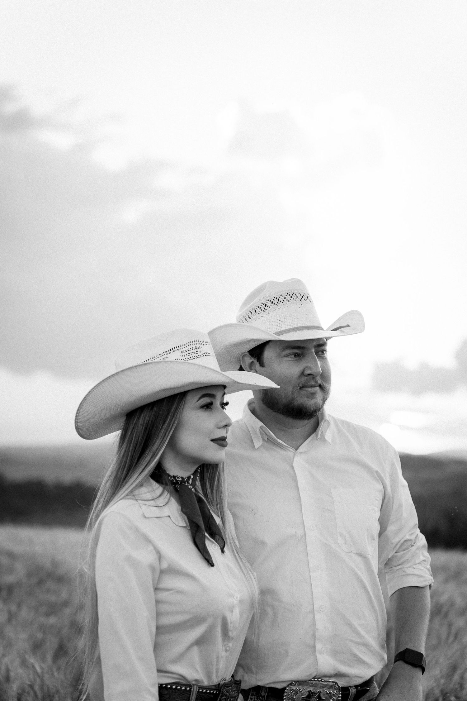 Black and white photo of a couple in cowboy hats and white shirts standing in a field at dusk.