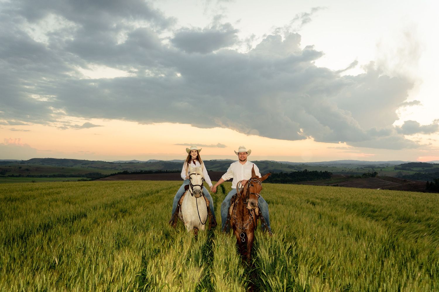 Couple riding horses through a lush green field at sunset wearing cowboy hats in a scenic rural landscape.