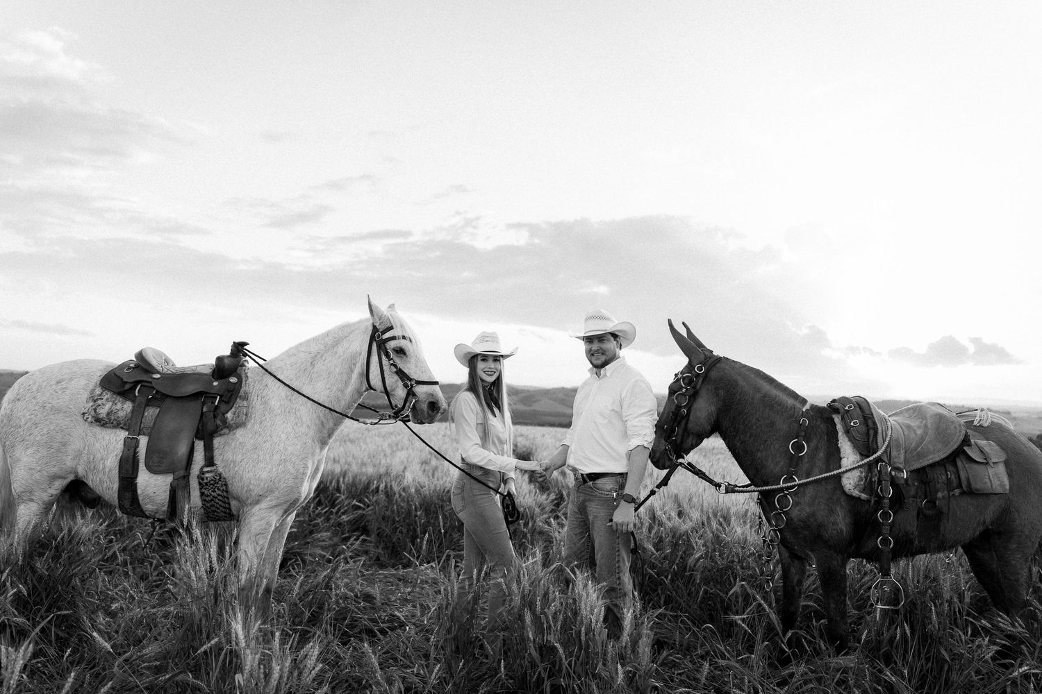 A couple in cowboy hats stands between two saddled horses in an open field in this black and white photo.