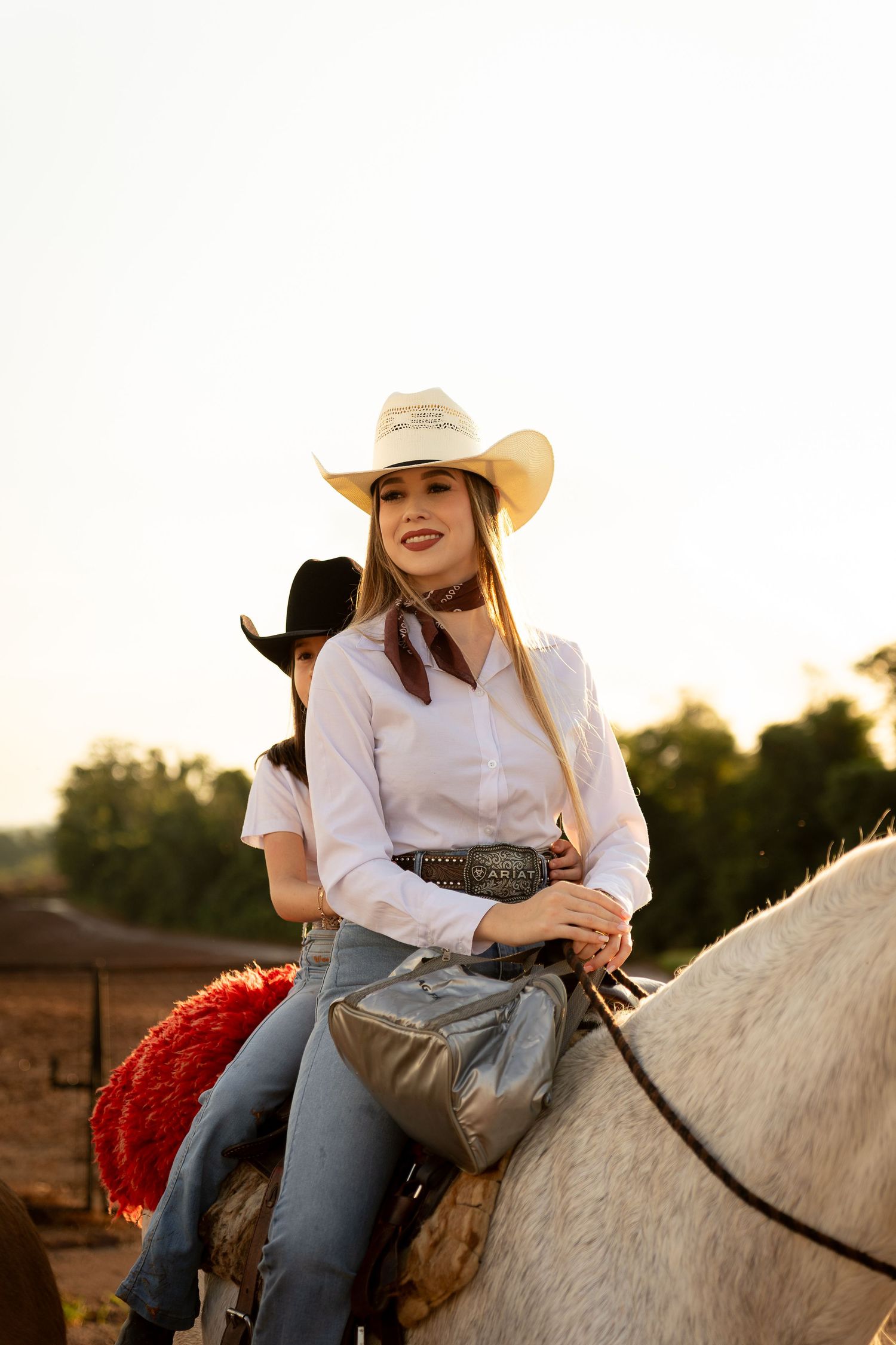Young woman in white cowboy hat and jeans riding a white horse at golden hour on a ranch.