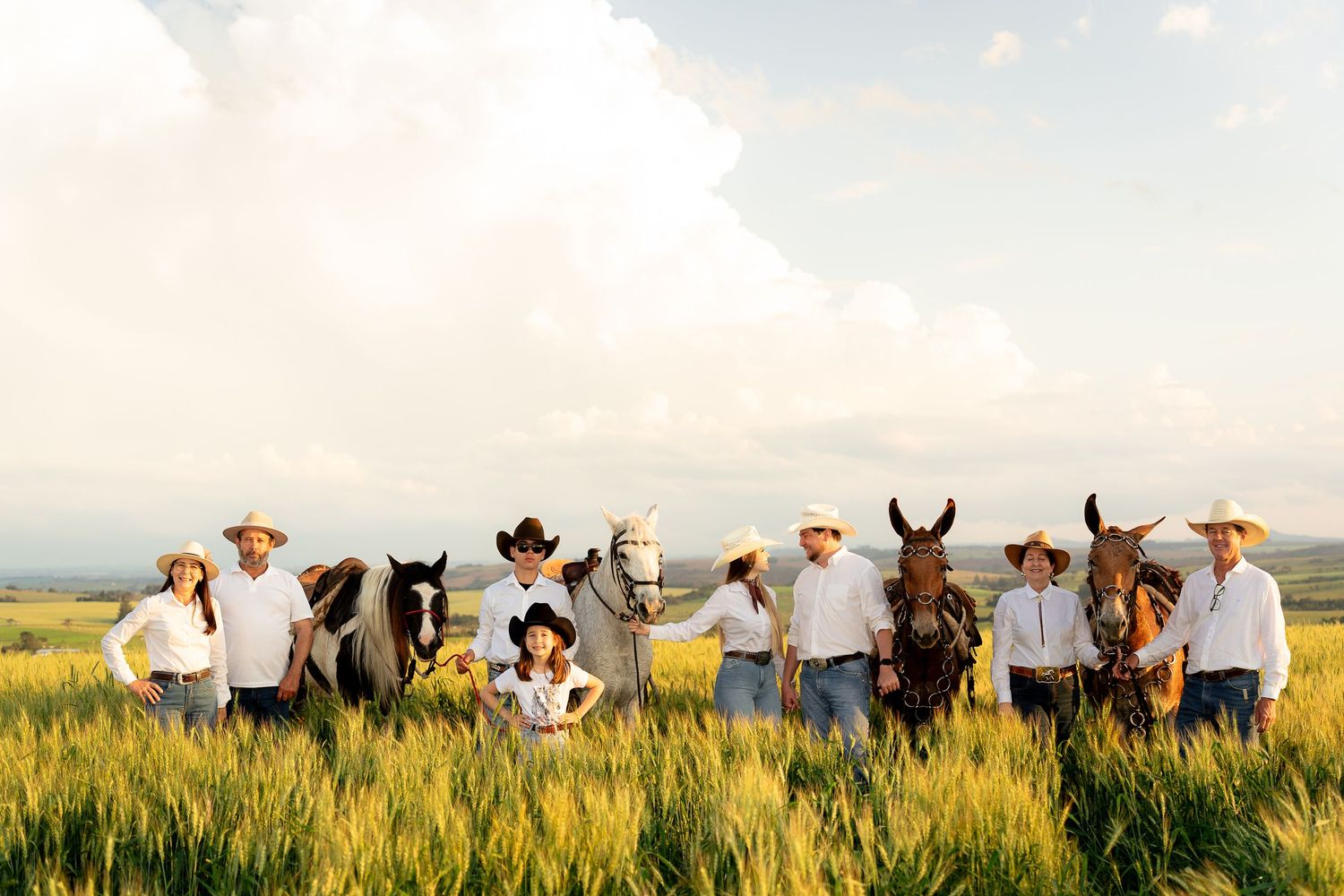 A group of cowboys and cowgirls in white hats stand with horses in a lush green field under a dramatic cloudy sky.