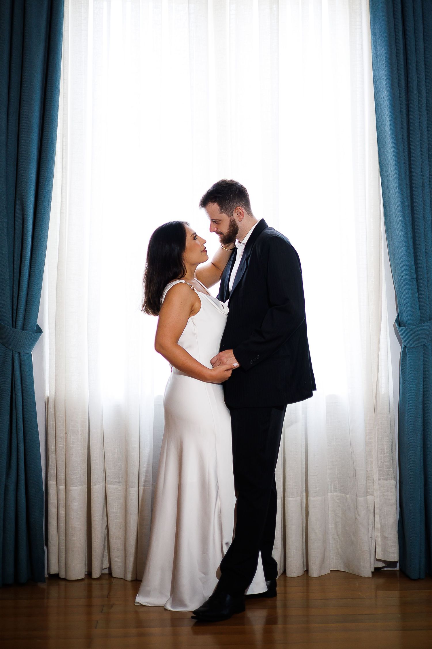 Elegant couple in wedding attire embracing by a bright window with teal curtains on their wedding day.