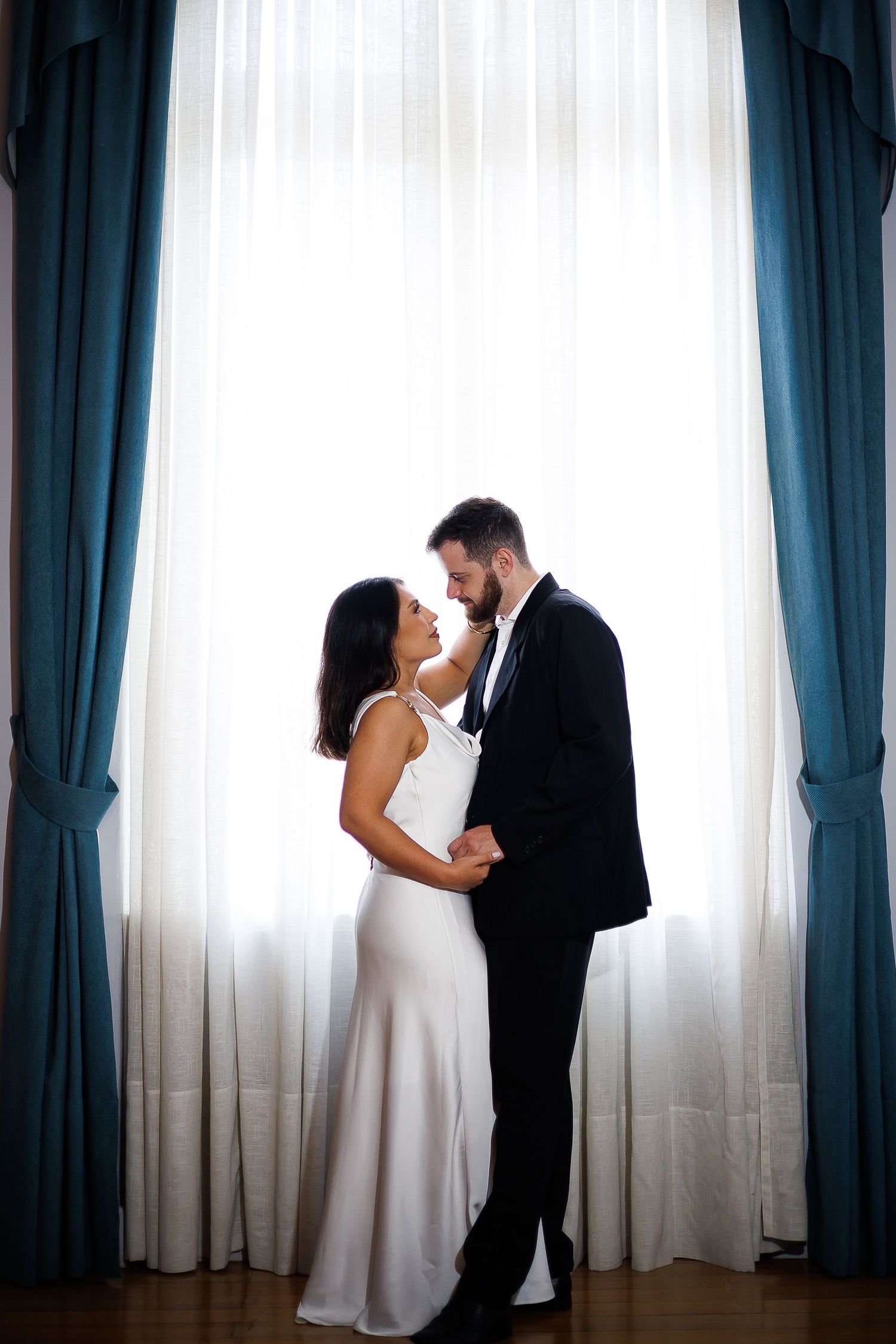 Elegant bride in white gown and groom in black suit embrace before large window with teal curtains.