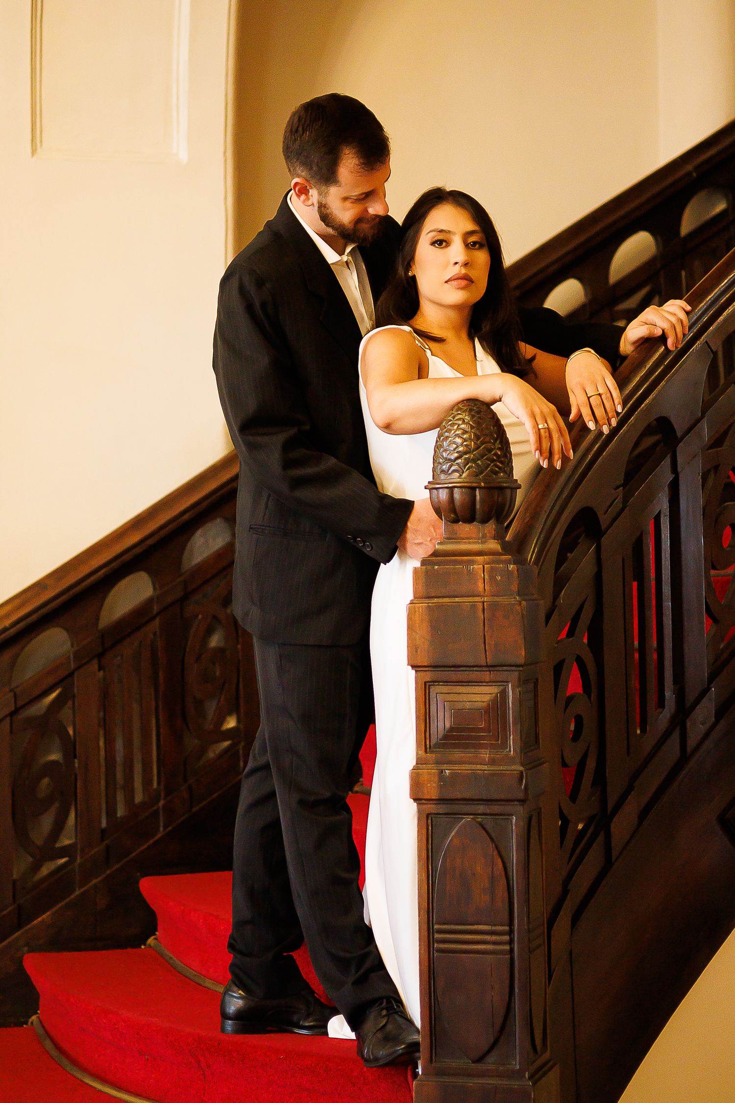 Elegant couple in formal attire posing on ornate wooden staircase with red carpet in a classic interior.