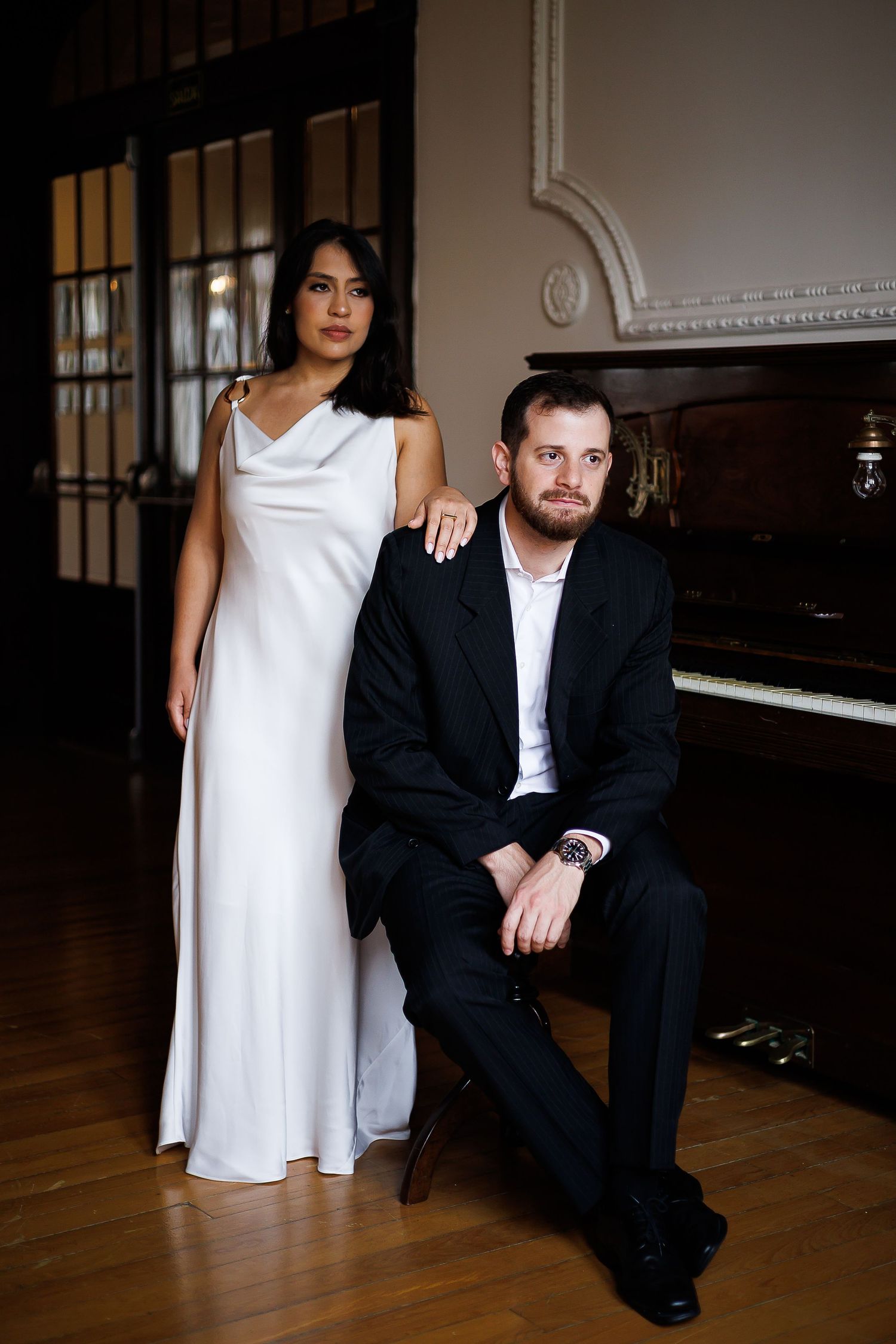 Elegant couple posing by a grand piano, woman in white gown, man in black suit, in a classic interior.