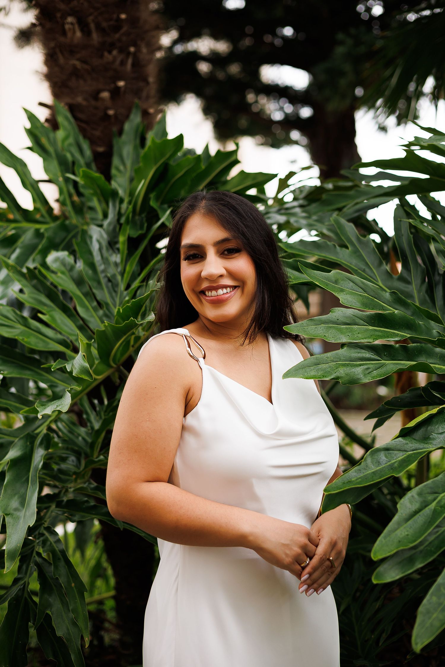 Smiling woman in white satin slip dress posing among tropical green leaves outdoors.