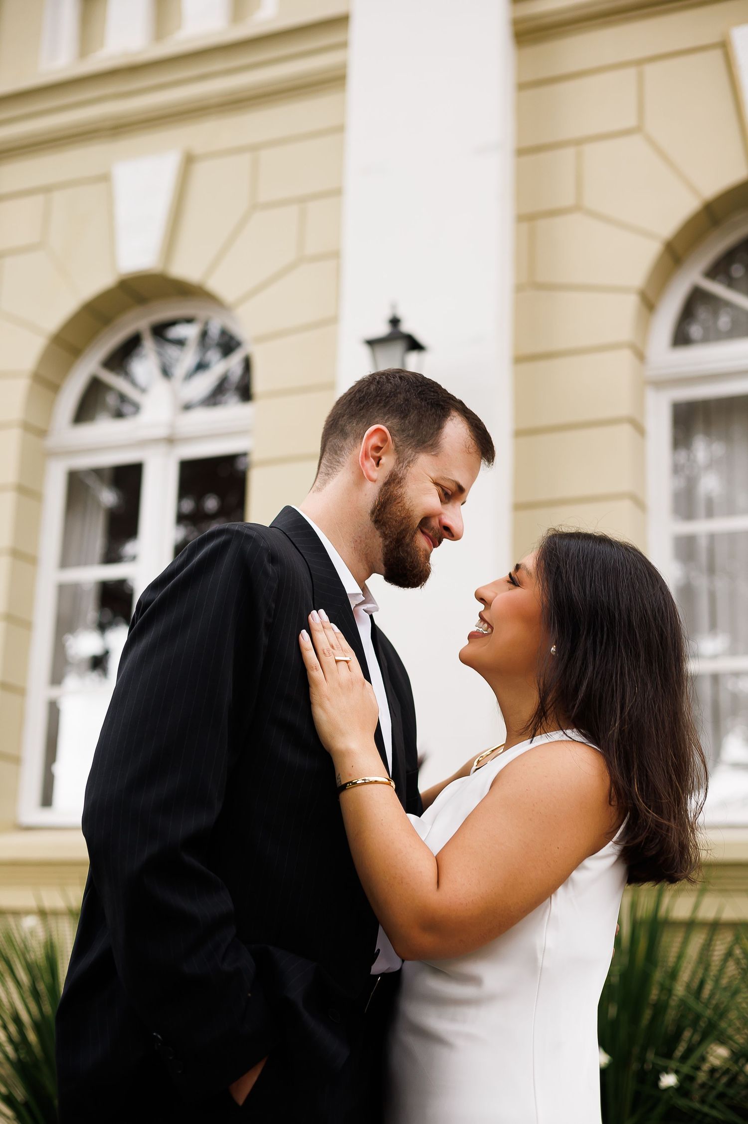 A couple in formal attire shares a romantic moment, smiling at each other in front of an elegant building.