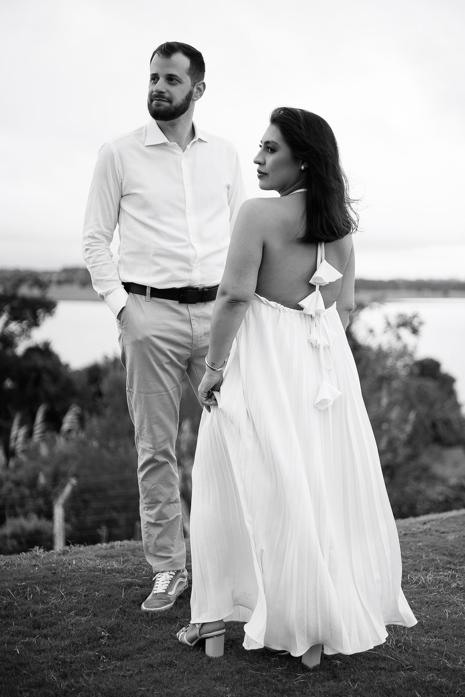 Black and white photo of an elegant couple outdoors, woman in backless white gown holding hands with man in white shirt.