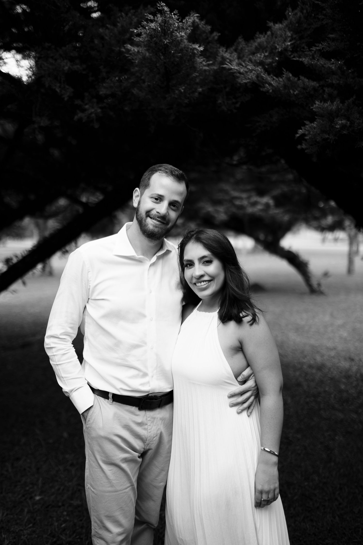 Black and white photo of a smiling couple posing outdoors under a large tree, man in white shirt, woman in white dress.
