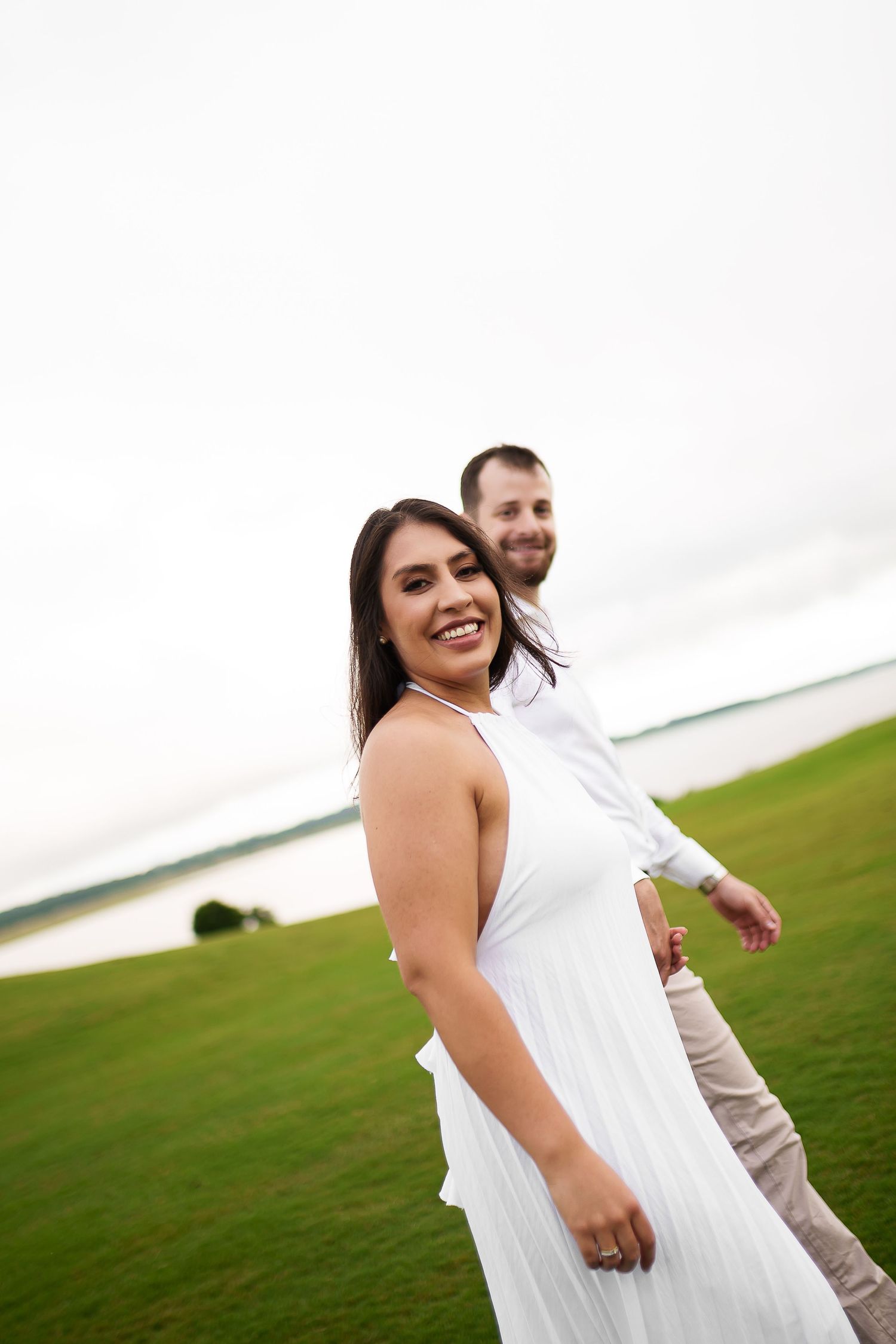 Smiling couple in white outfits on green lawn near water, woman in white dress laughing as man holds her hand.