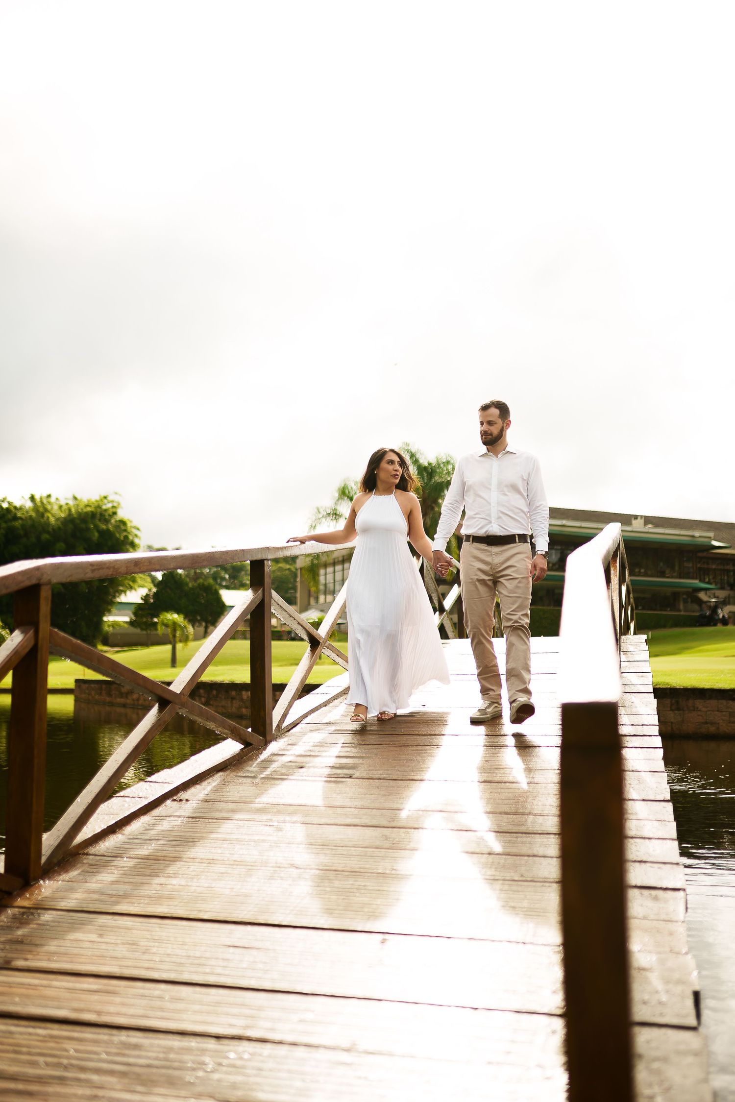 Couple walking hand in hand on a wooden bridge, woman in white dress, man in casual attire, lush green background.