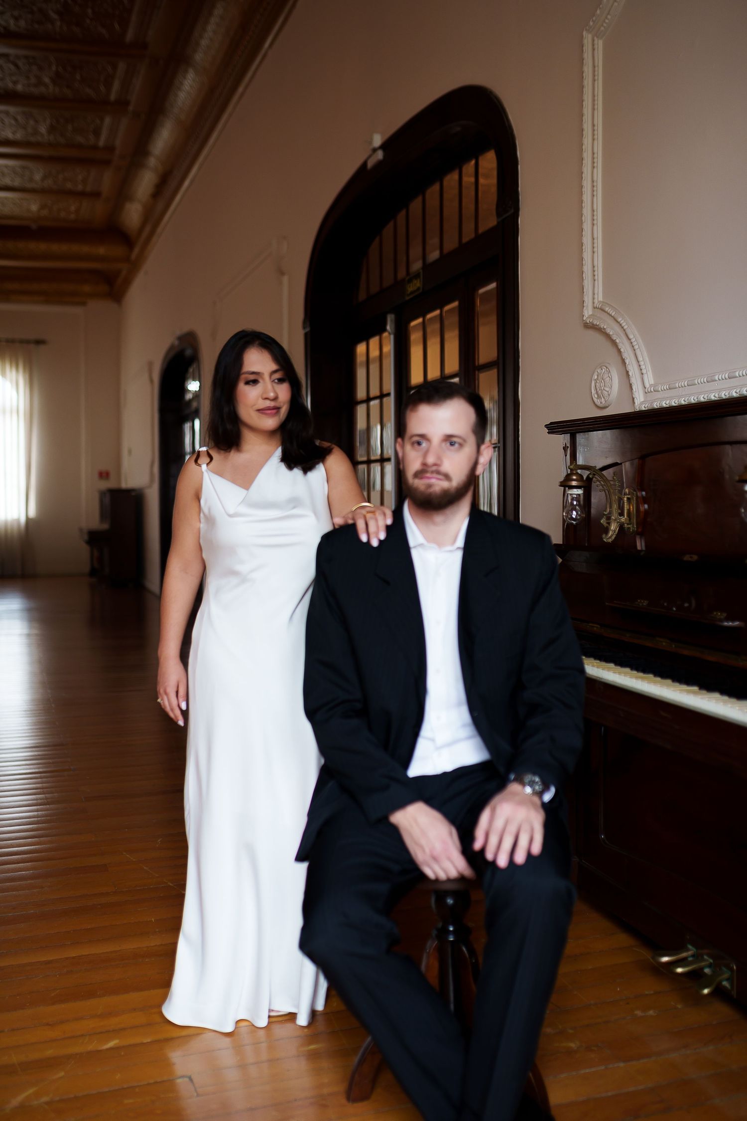 Couple in elegant attire poses near a vintage piano in a grand hall, woman in white gown, man in black suit seated.