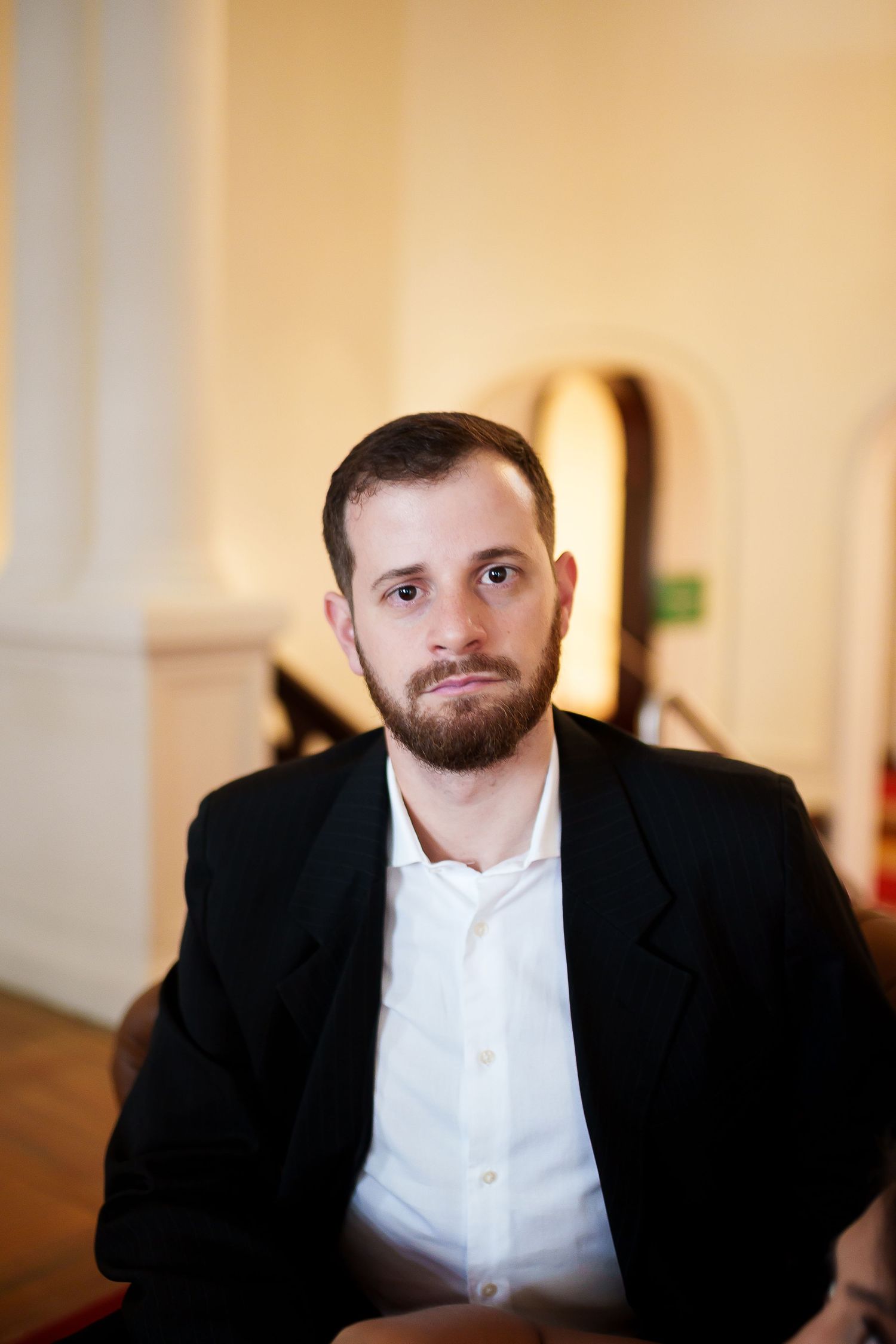 Bearded man in black blazer and white shirt sitting indoors with a neutral expression.