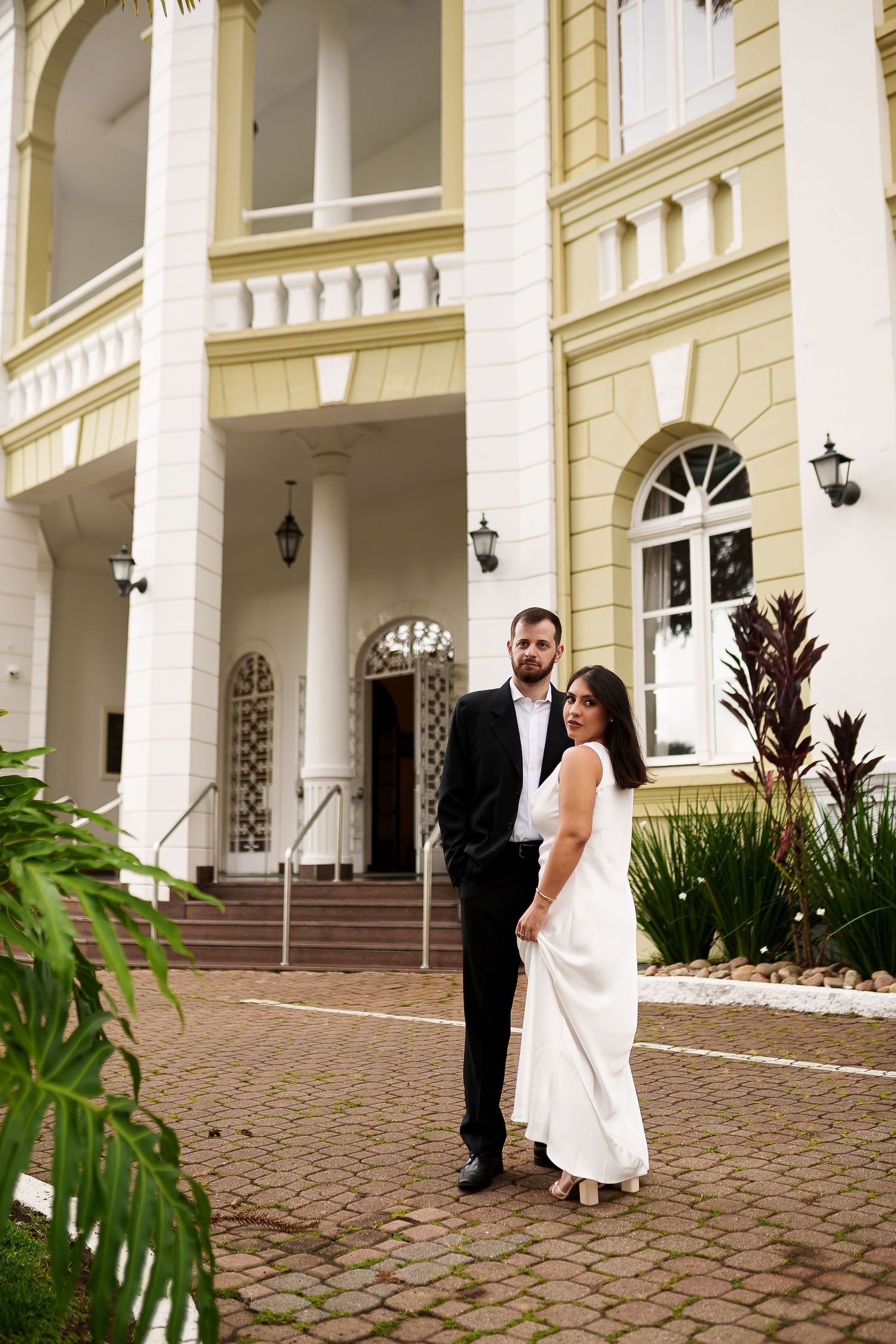 Elegant couple in wedding attire posing in front of a grand colonial-style building with arched windows.