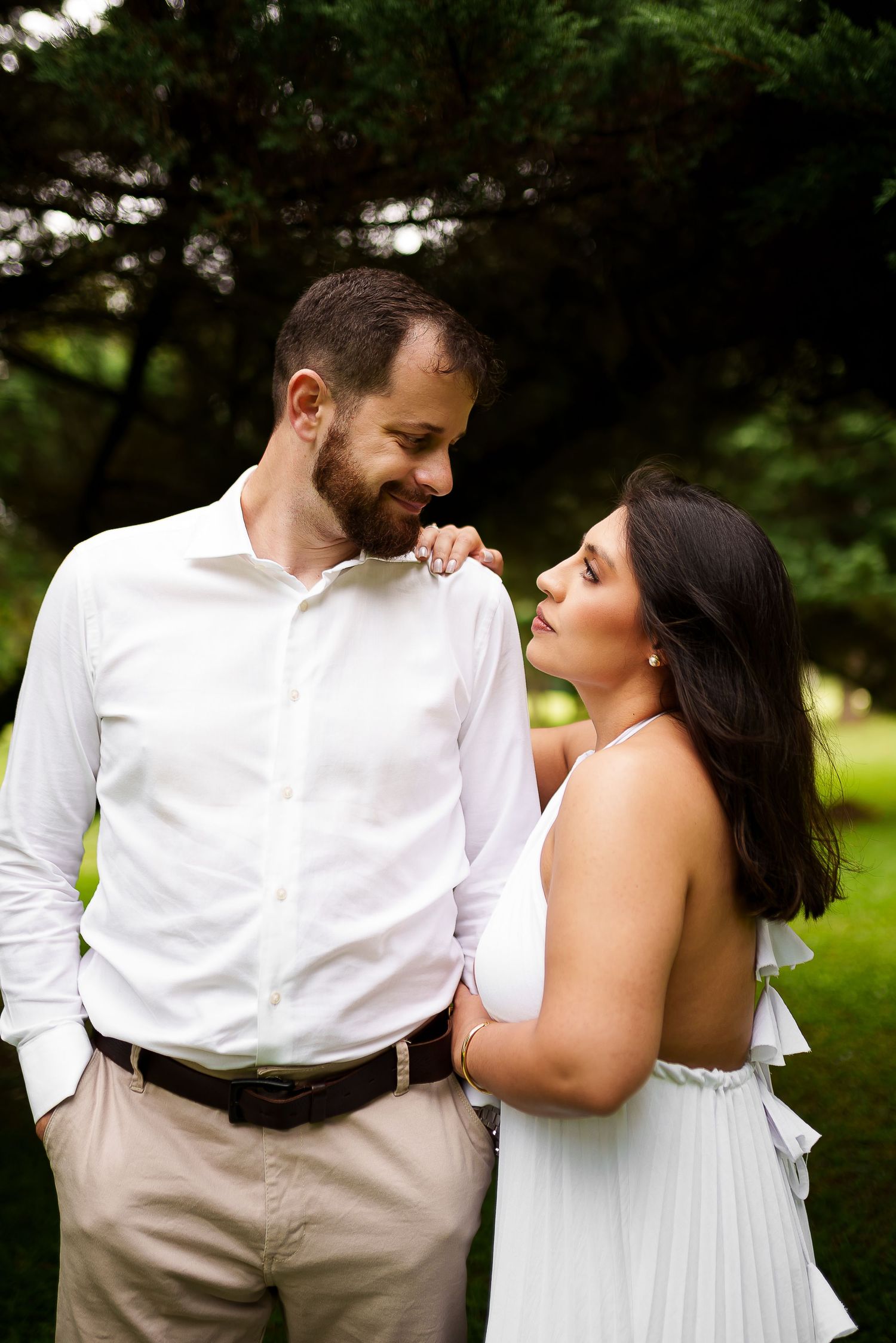 A couple in white outfits share a tender moment outdoors, smiling at each other in a lush green park setting.