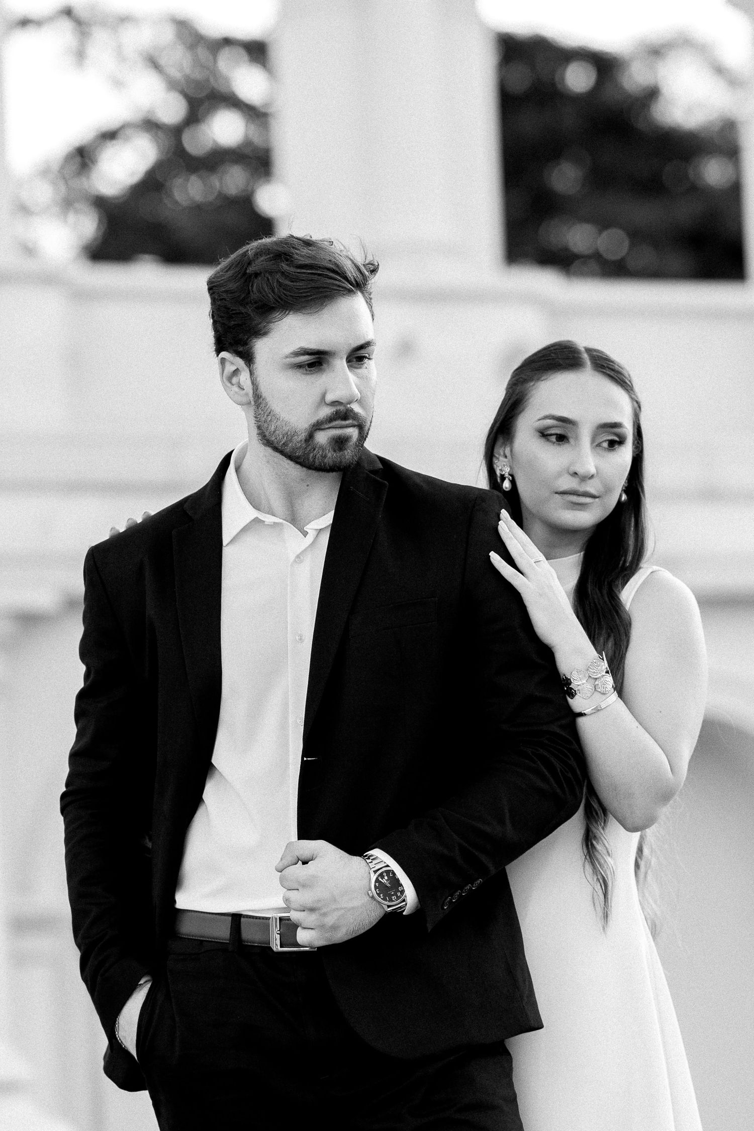 Black and white photo of an elegant couple, man in suit and woman in white dress, posing outdoors near columns.