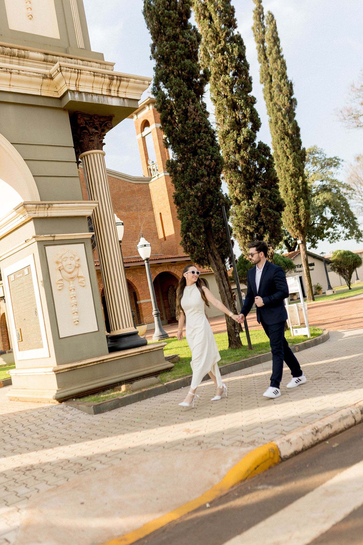 Couple holding hands walking near a historic church with tall cypress trees on a sunny day.