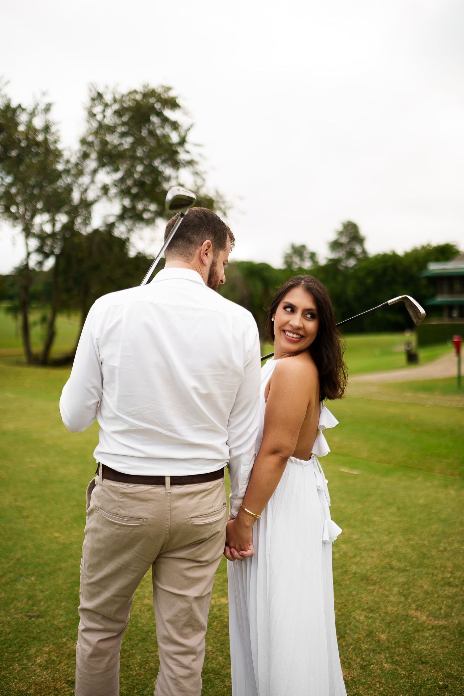 Couple holding hands on a golf course, woman in white dress smiling over shoulder, man in white shirt holding golf club.