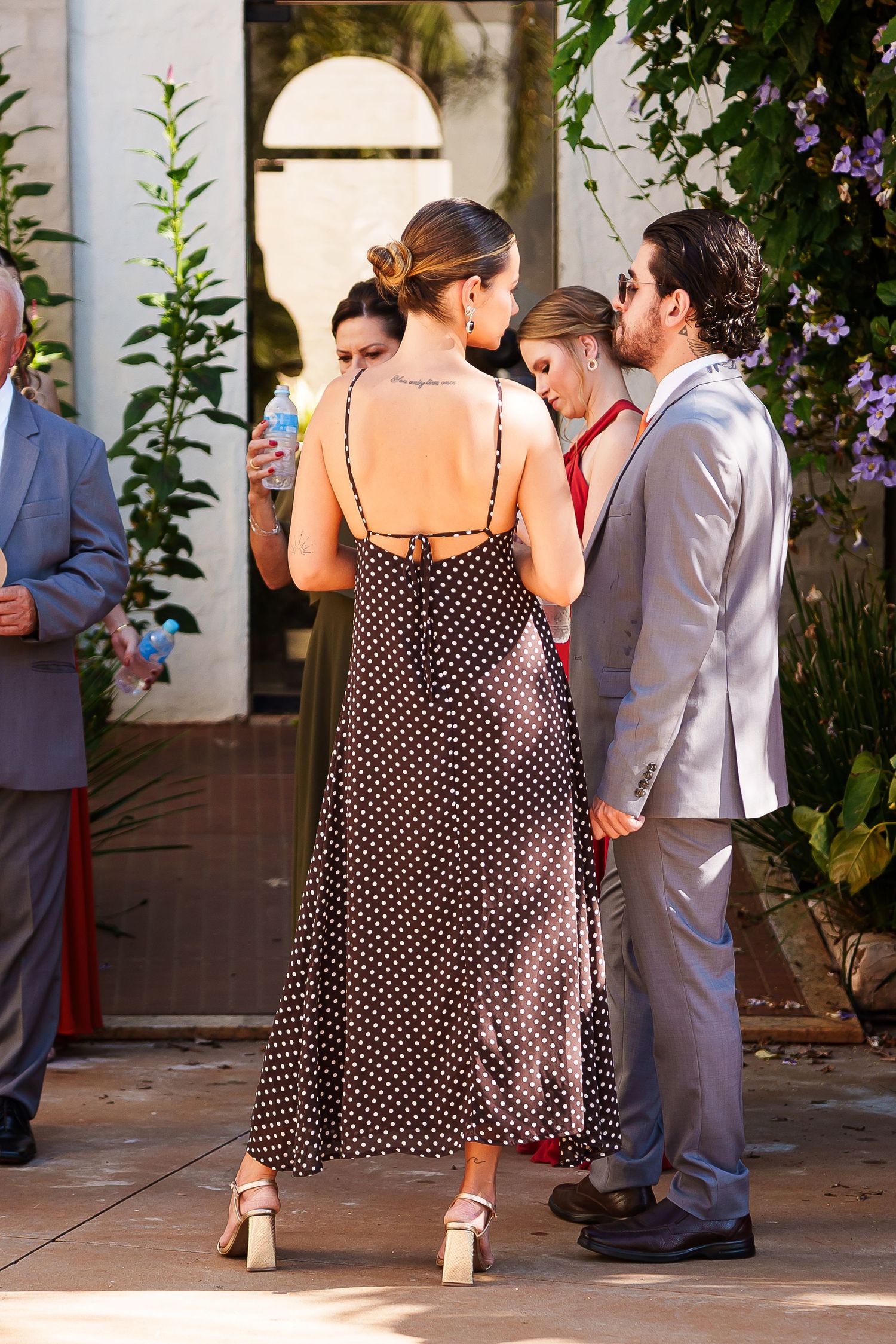Couple in polka dot dress and gray suit at outdoor garden event with floral backdrop.