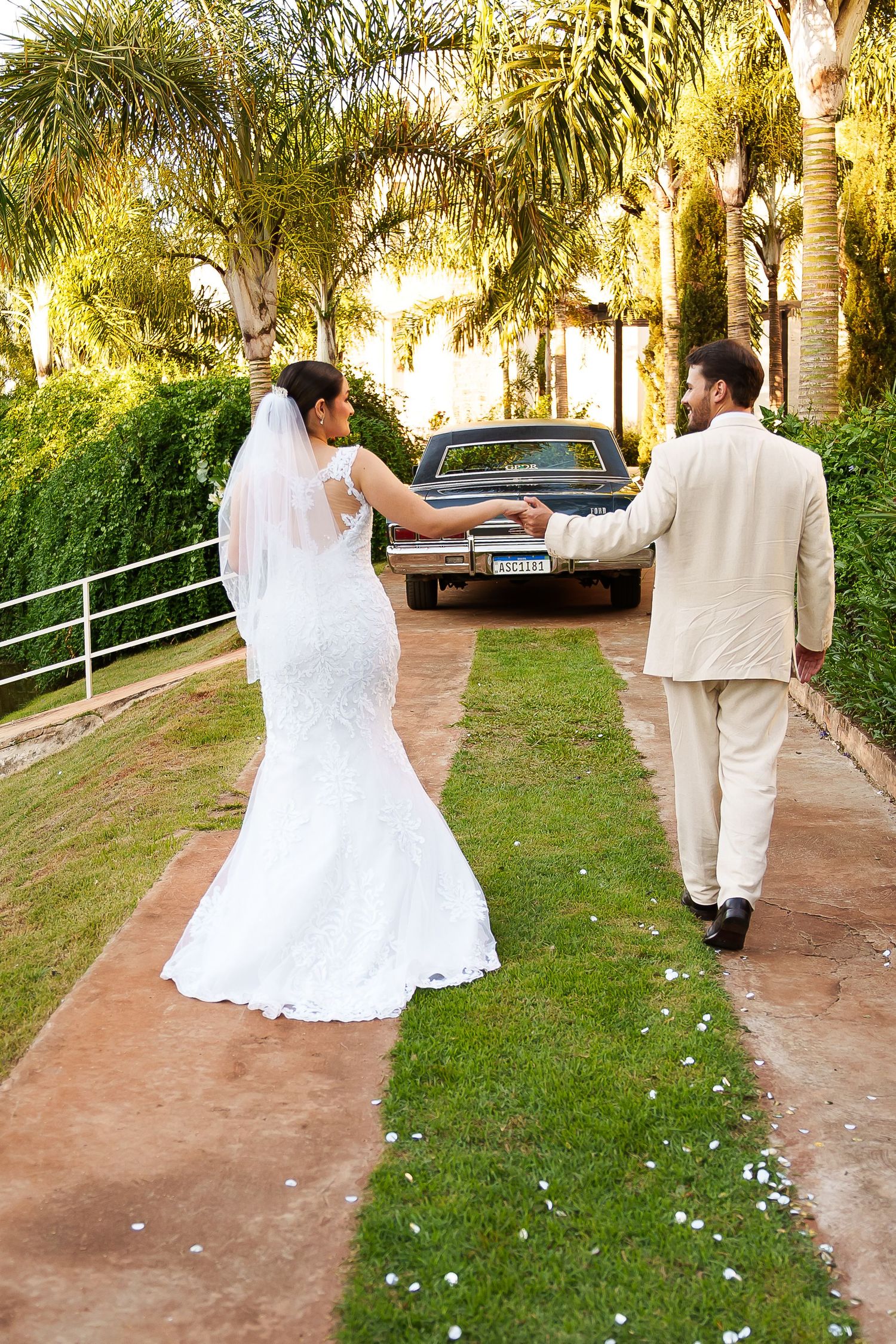 Bride and groom walking hand in hand toward a vintage car along a palm-lined path on their wedding day.