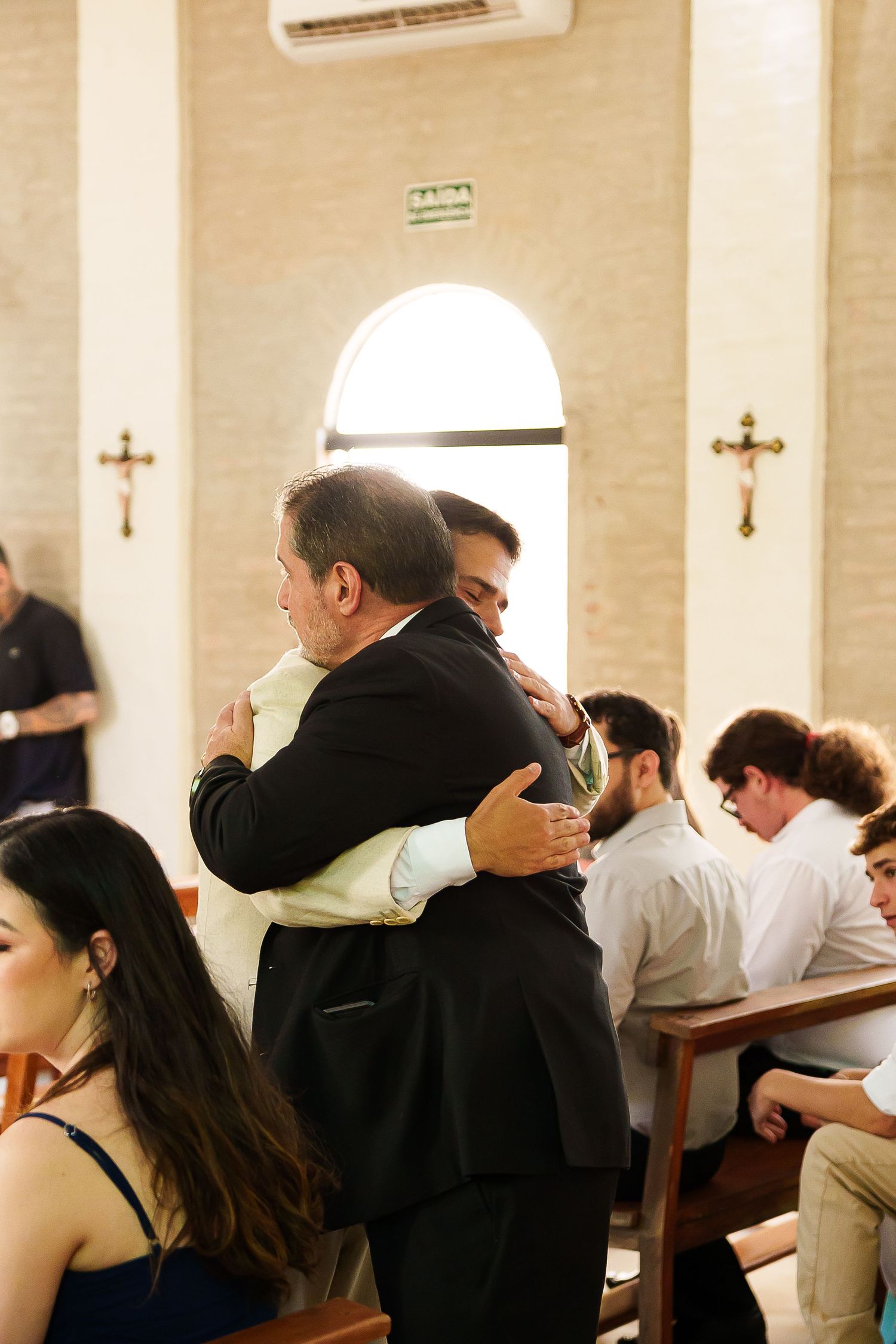 Two men share a warm embrace inside a church during a ceremony, with guests seated in pews and crosses on the walls.