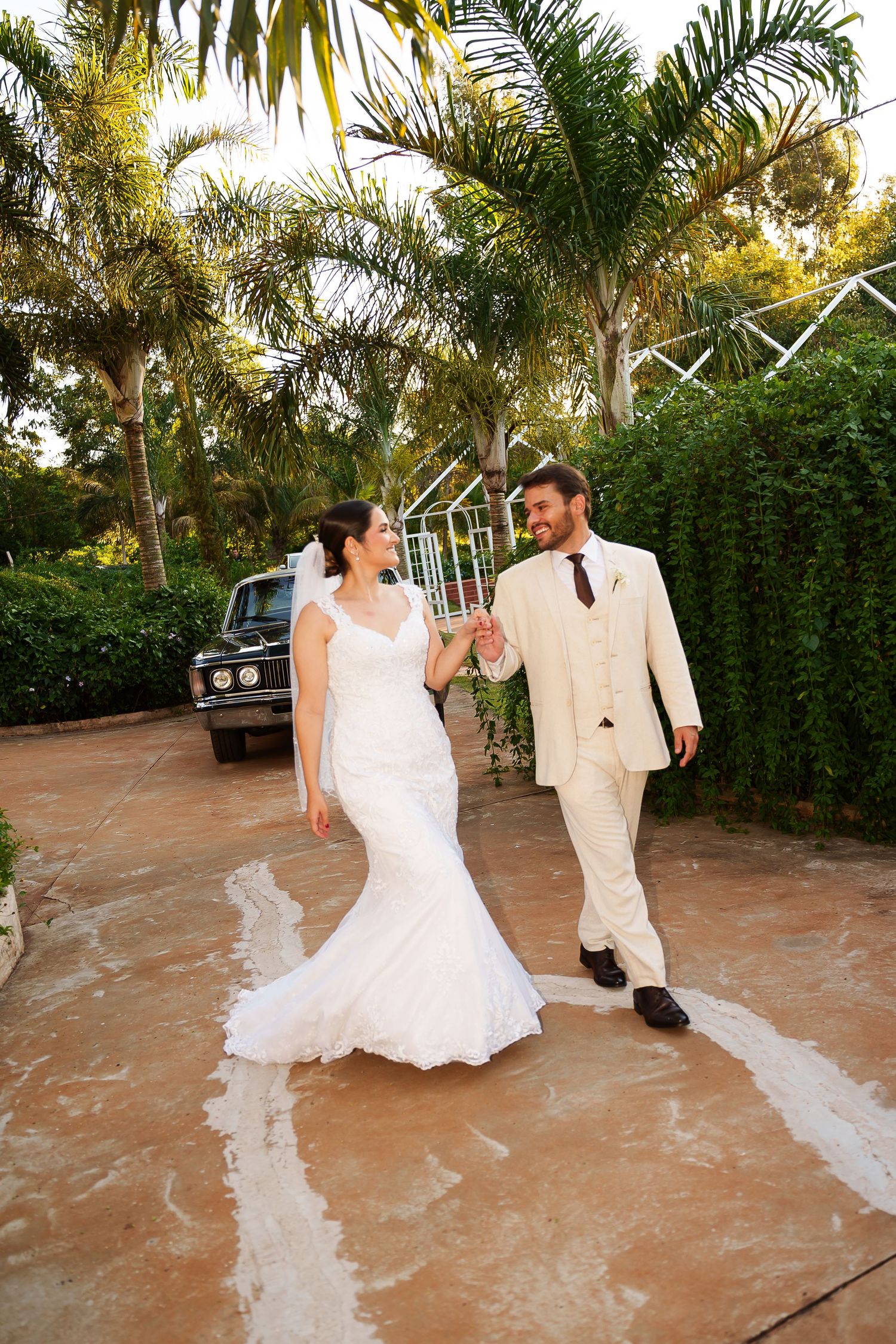 Bride in white mermaid gown and groom in beige suit hold hands walking outdoors near palm trees on their wedding day.