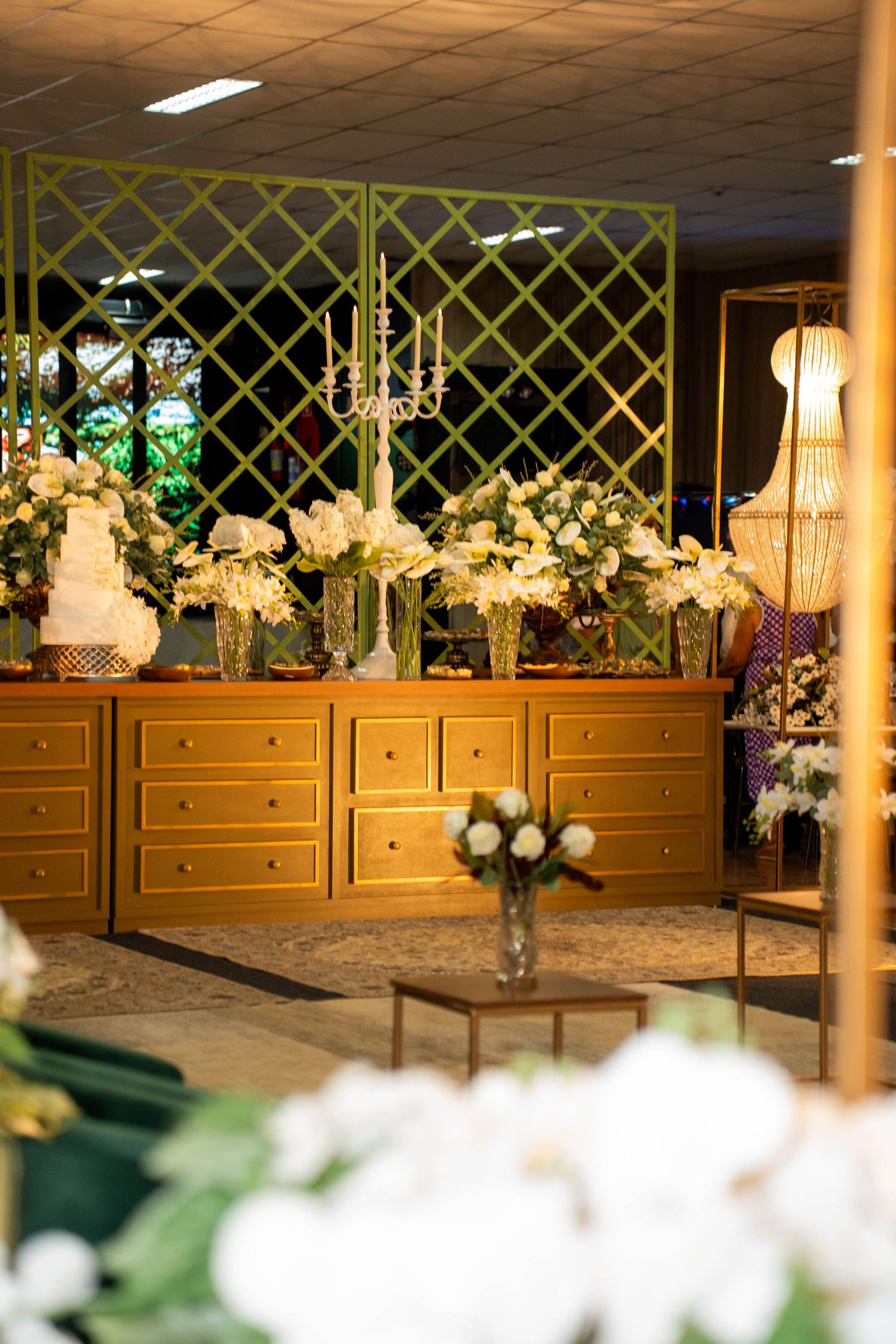 Elegant wedding reception display with white floral arrangements, candelabra, and tiered cake on golden dresser.