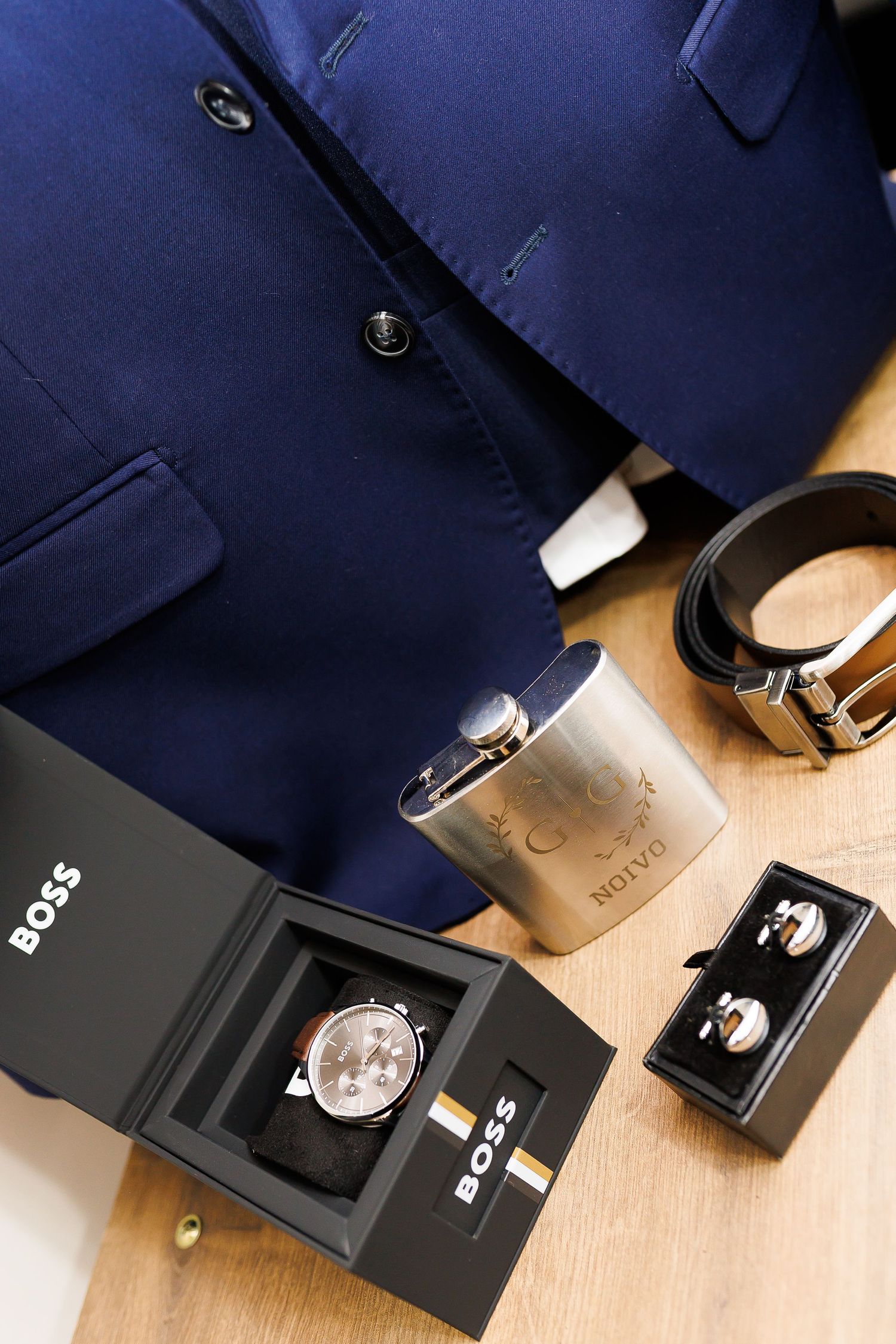 Navy blue suit with Hugo Boss watch, flask, cufflinks, and leather belt arranged on wooden surface.
